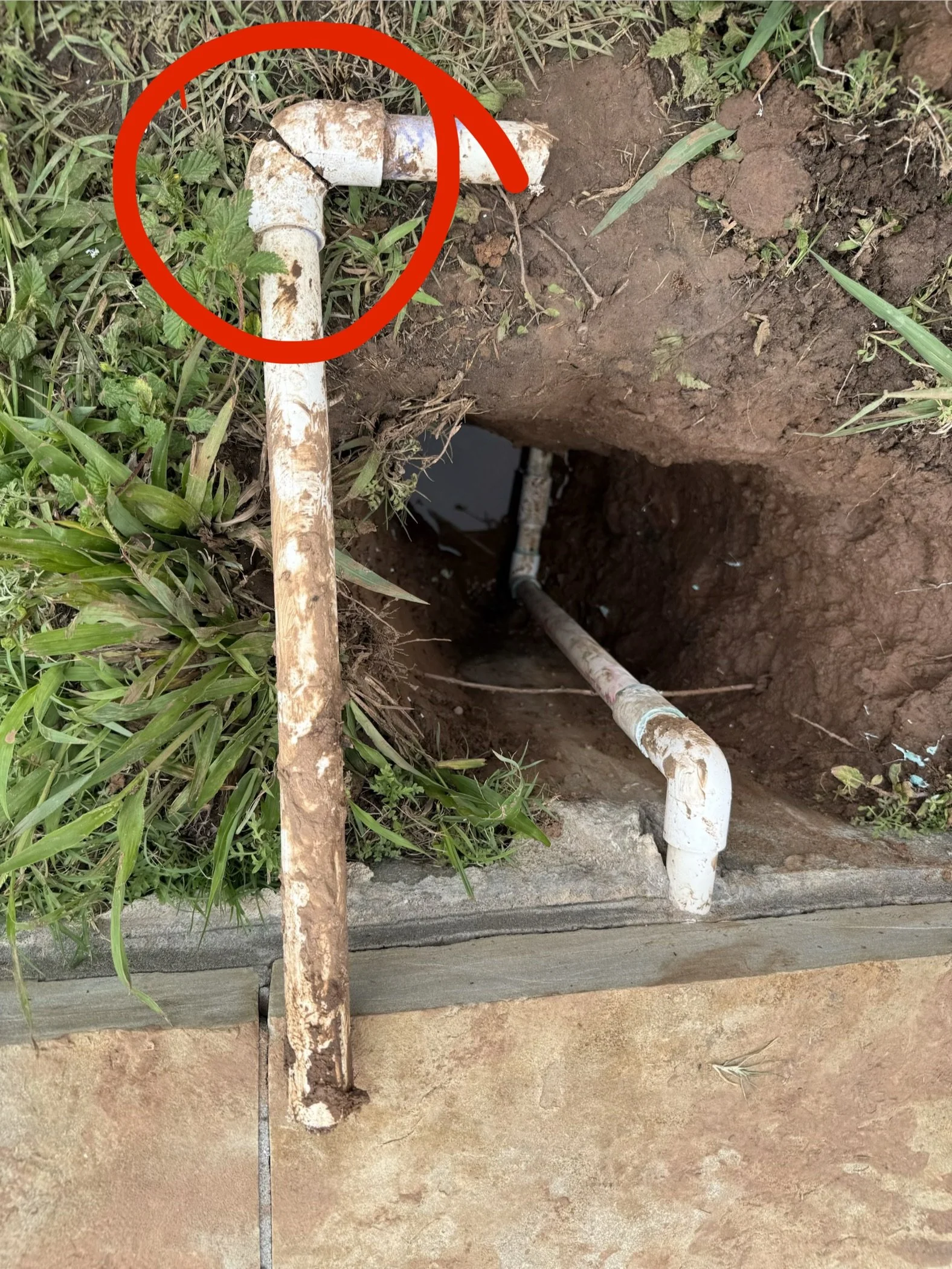 A rusty pipe protruding from the ground in a grassy area with a red circle and slash mark drawn over it, indicating a broken or leaking pipe.