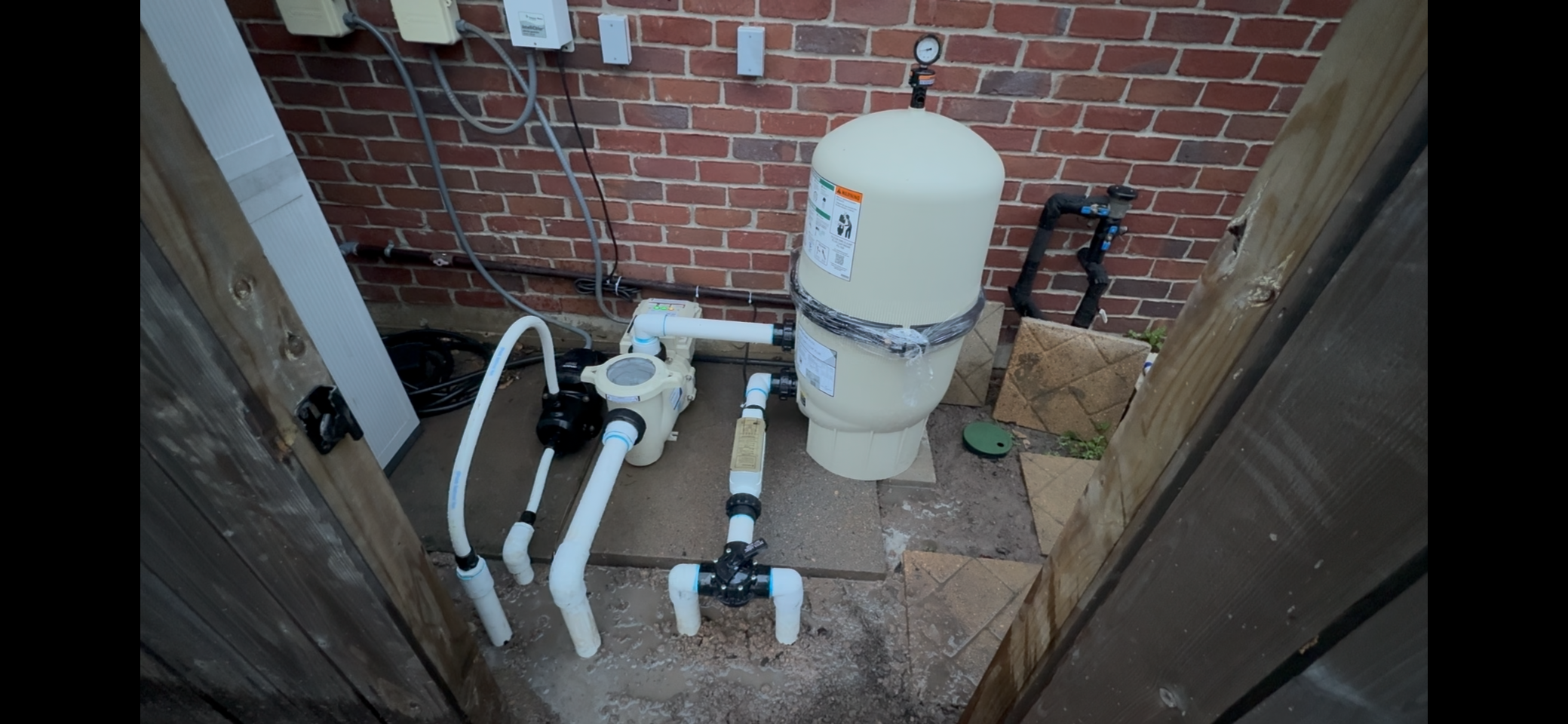 A pool filtration system with white pipes, a pump, and a filter in an outdoor area next to a brick wall, with some electrical wiring and a small patch of soil with bricks and grass in the corner.