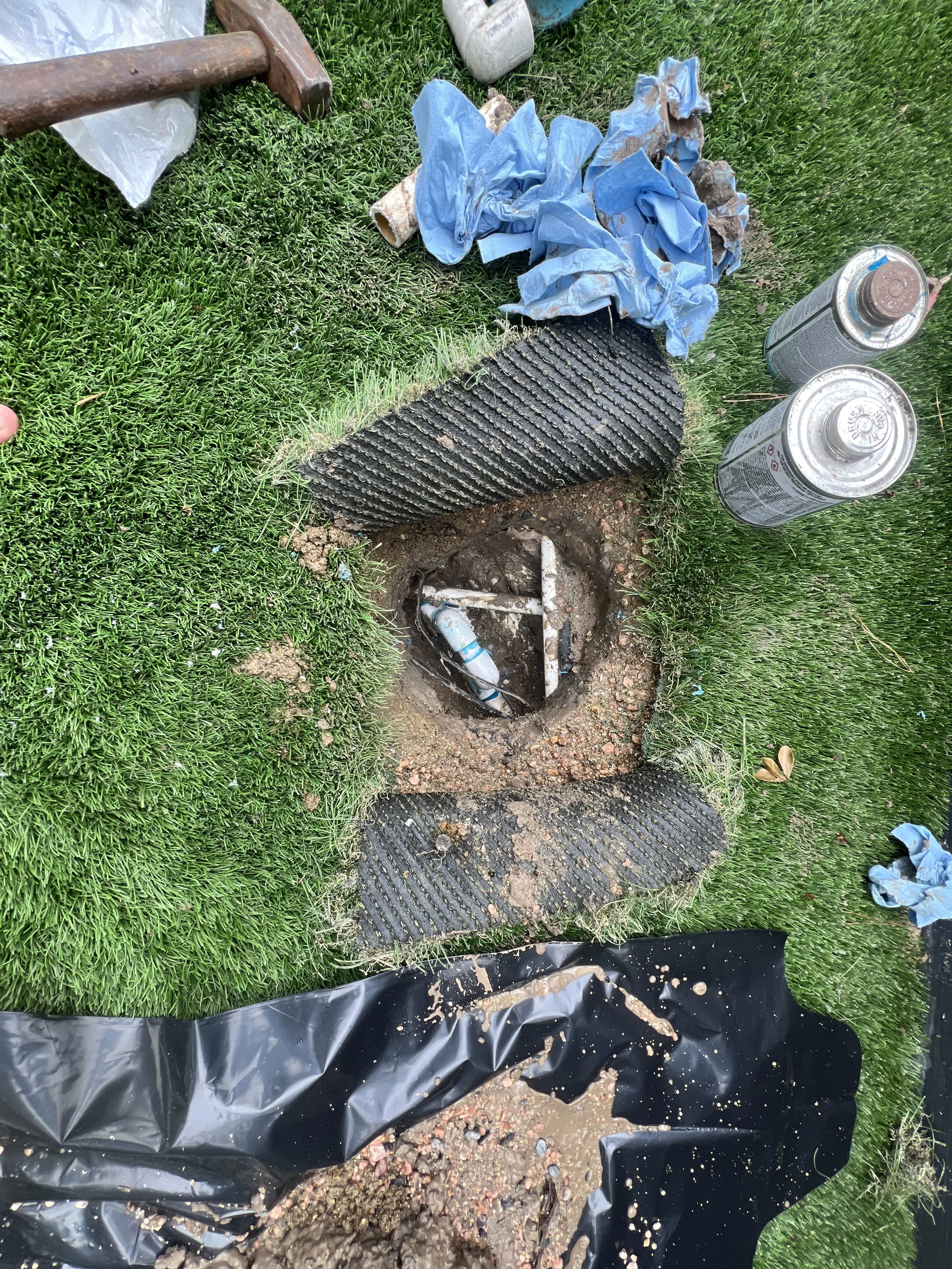 A trench filled with water, gravel, and soil, with pipes and plumbing visible. Surrounding it is grassy ground and construction tools, including spray cans, gloves, and a blue rag.
