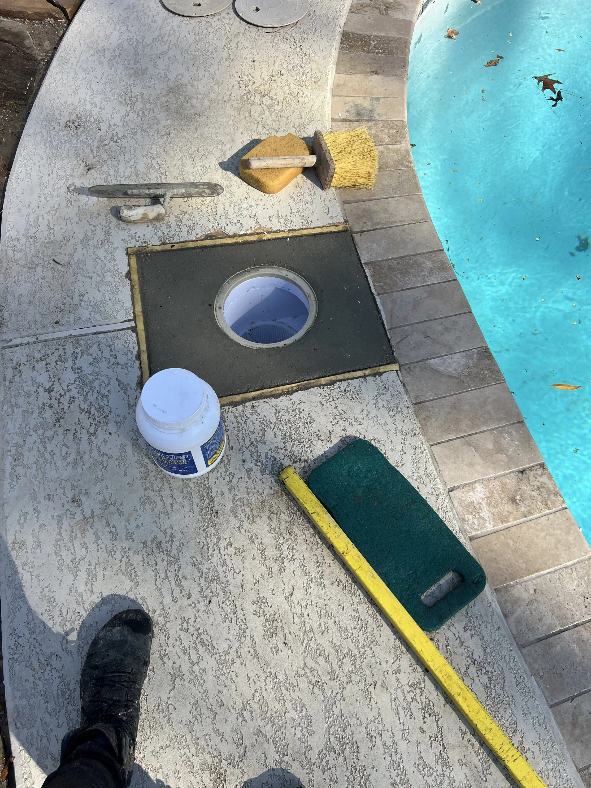A poolside area with cleaning tools including a yellow broom, a yellow sponge, a scrub brush, and a bucket of cleaning solution. The person’s black work shoes are visible in the bottom left corner, and a yellow level tool is resting on a green mat. T