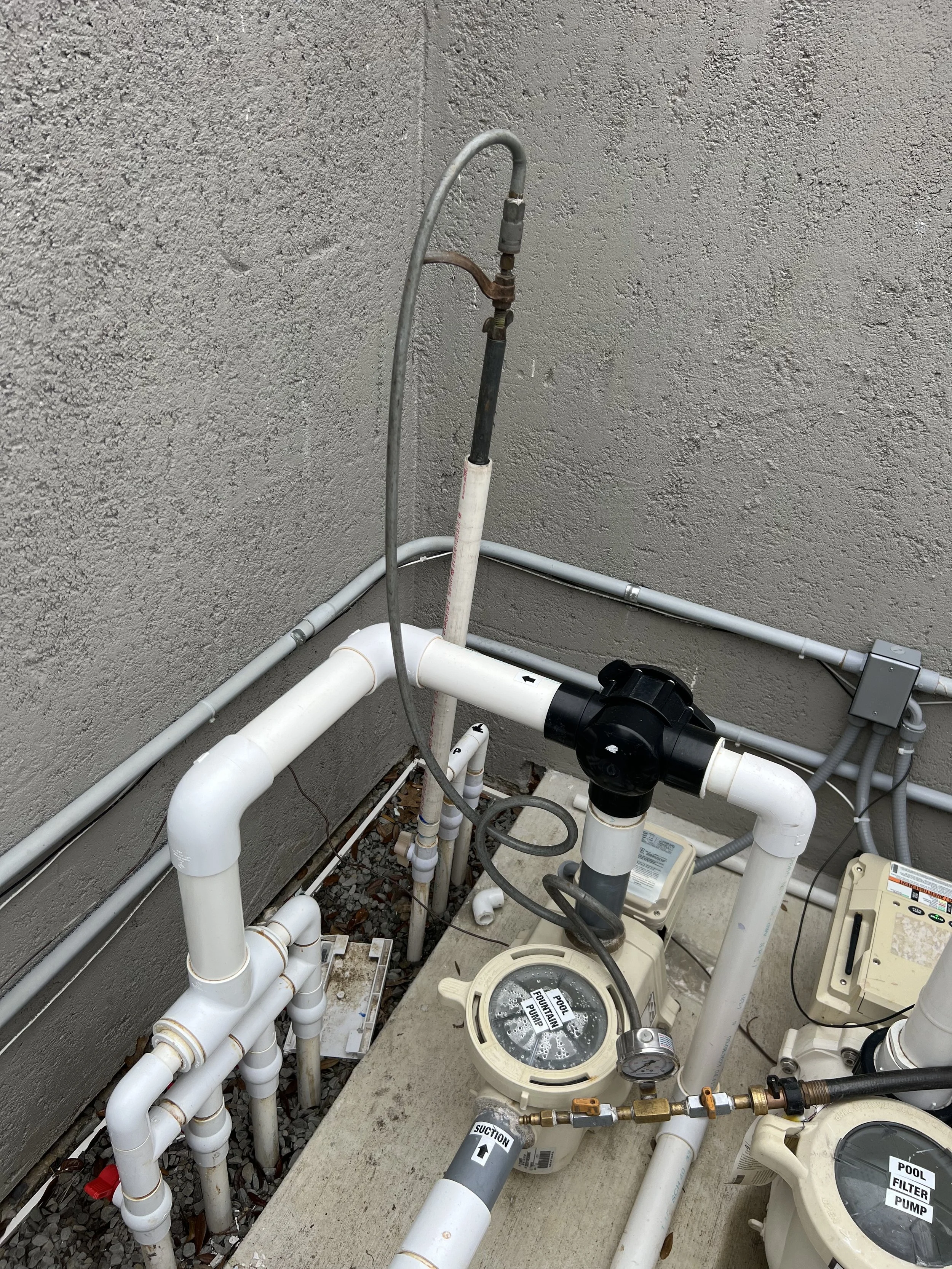 Various plumbing pipes, valves, filters, and pumps installed outside on a concrete surface against a textured wall.