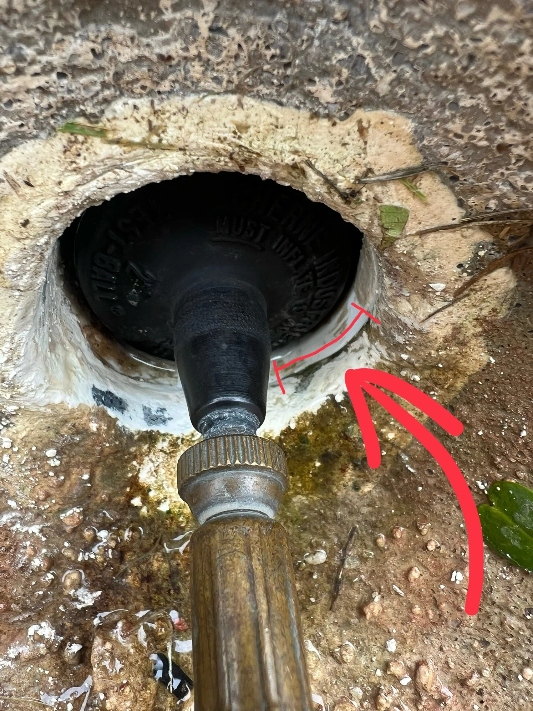Close-up of a pipe installed in a concrete foundation with a red arrow pointing to it, surrounded by dirt and small rocks.