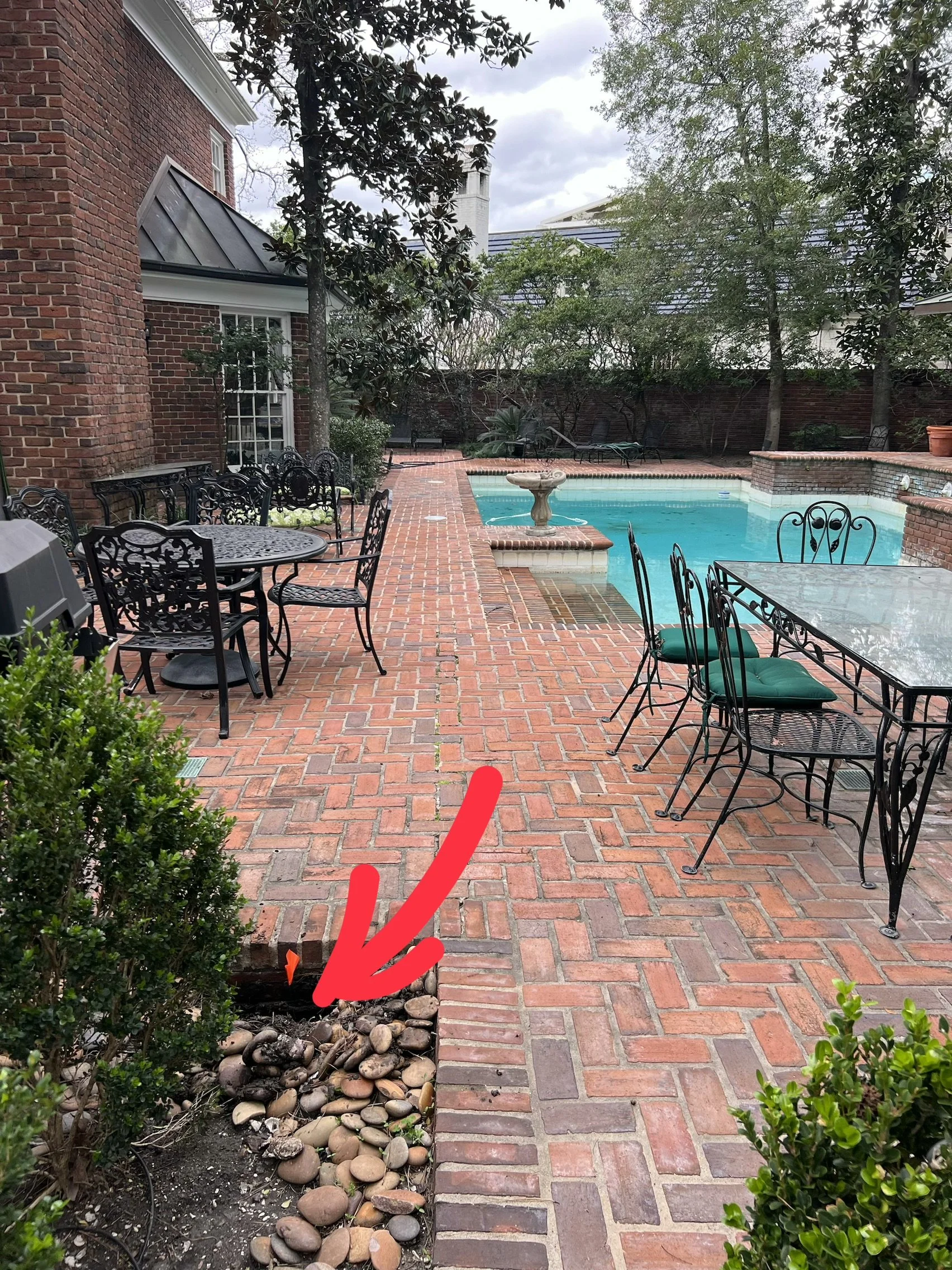 A brick patio with outdoor tables and chairs, a swimming pool, and garden plants. A small trench with rocks and dirt is visible next to the patio with a red arrow pointing to it.