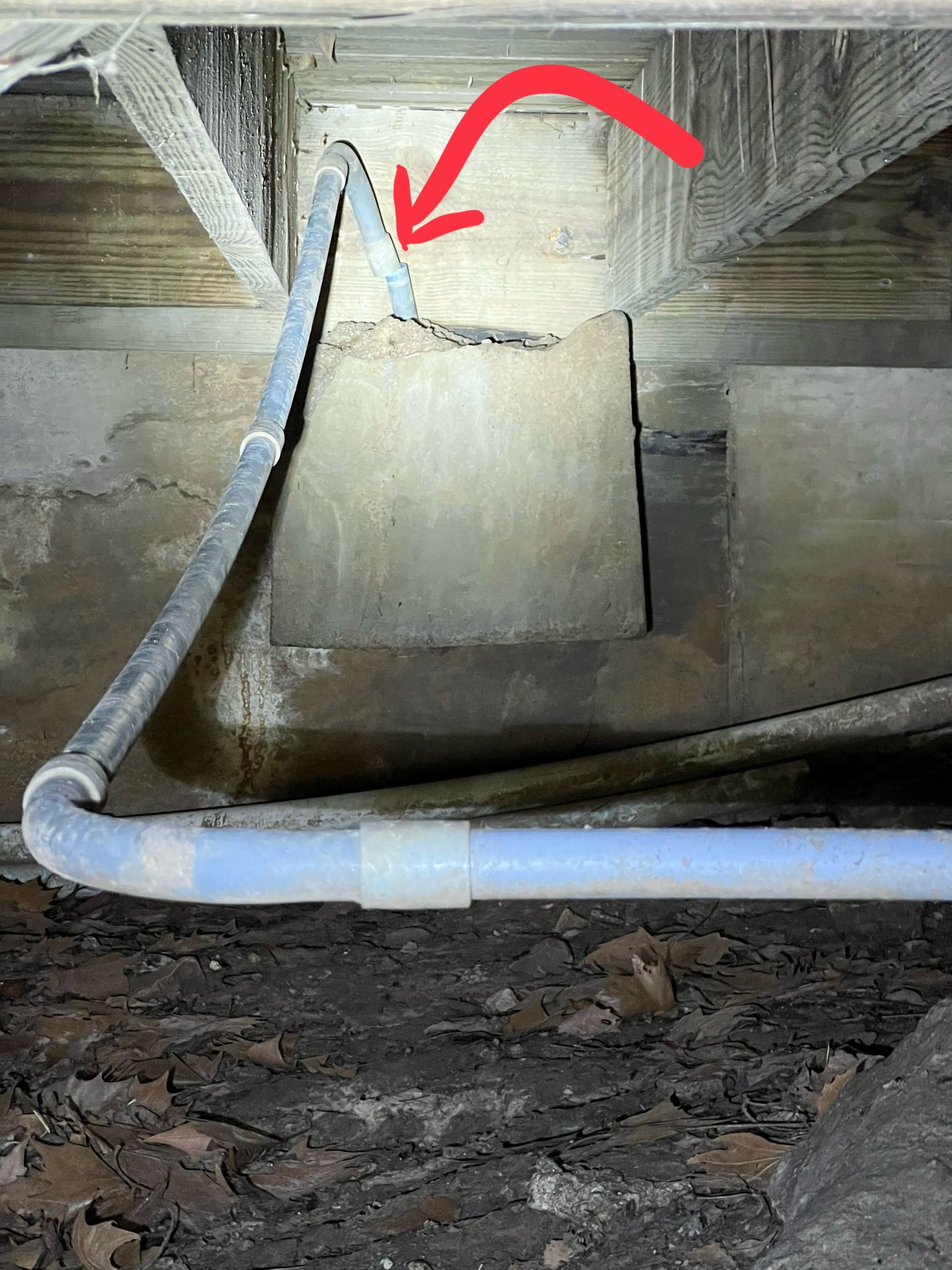View of a crawl space or underfloor area with exposed pipes, wooden framing, and dirt ground, highlighted by a red arrow.