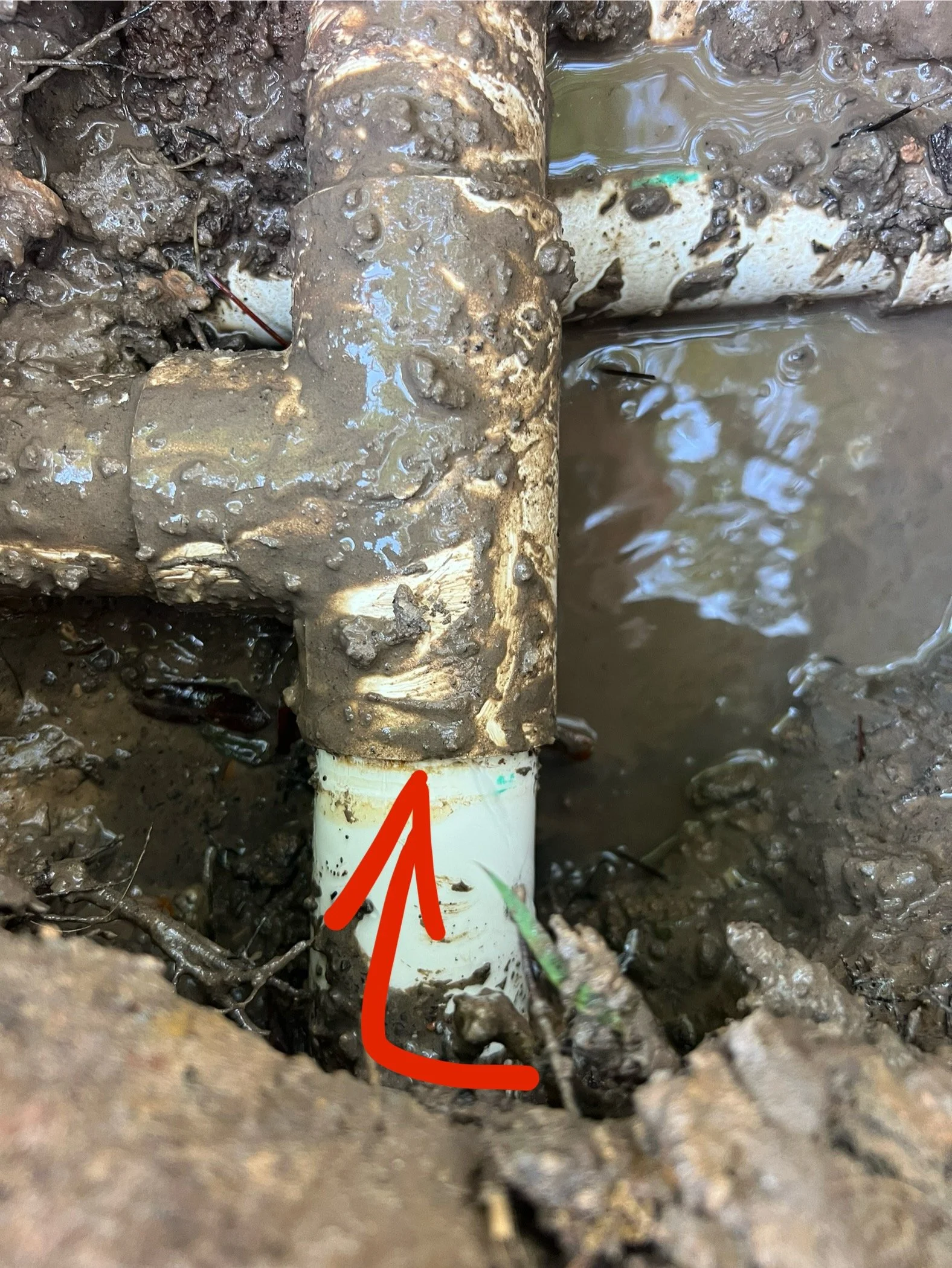Close-up of a white PVC pipe partially buried in muddy soil, with a rusty metal pipe connected to it, surrounded by wet soil, and marked with a red arrow.