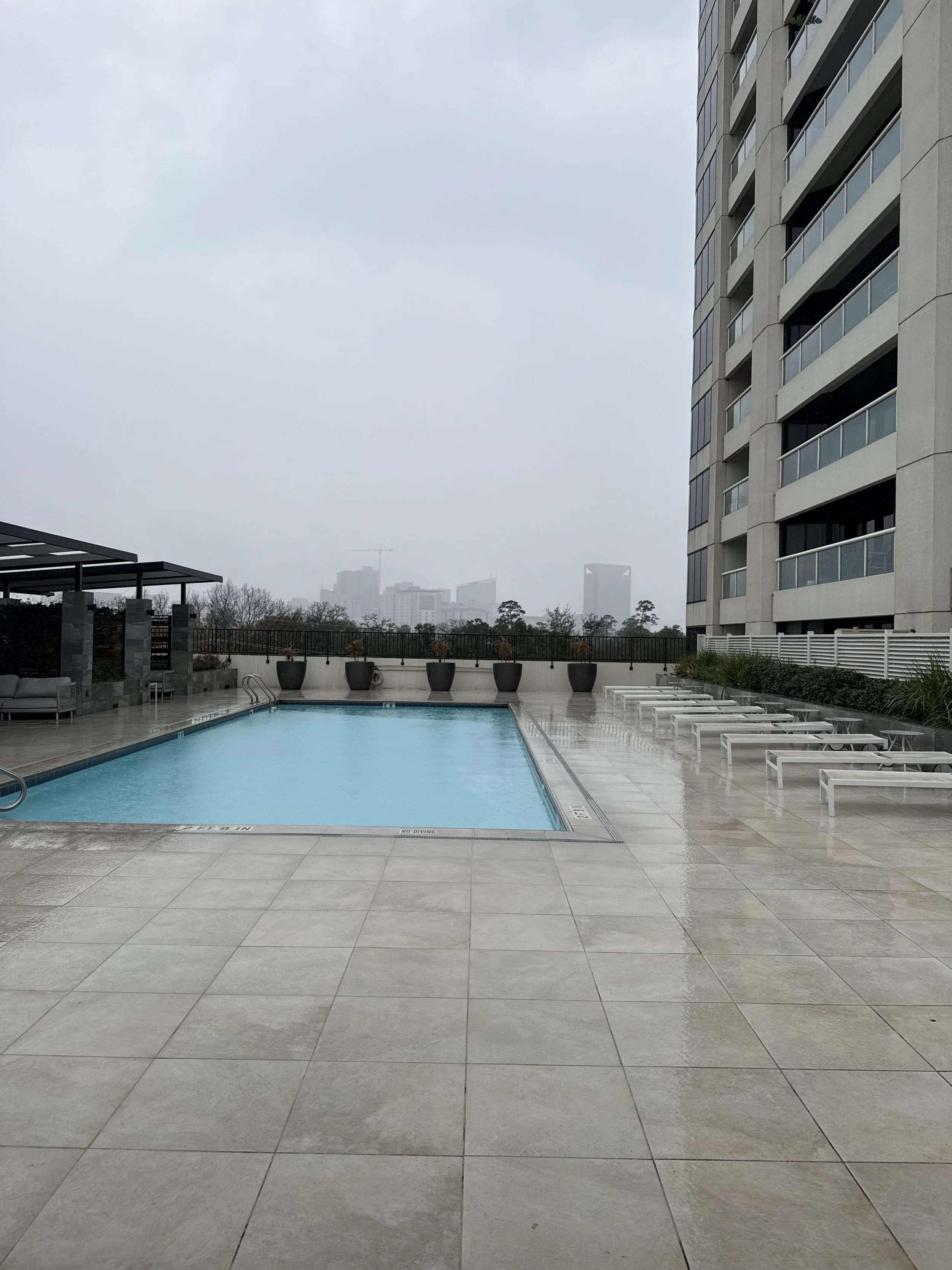 An outdoor swimming pool area with lounge chairs and potted plants, surrounded by modern high-rise buildings on a cloudy day.