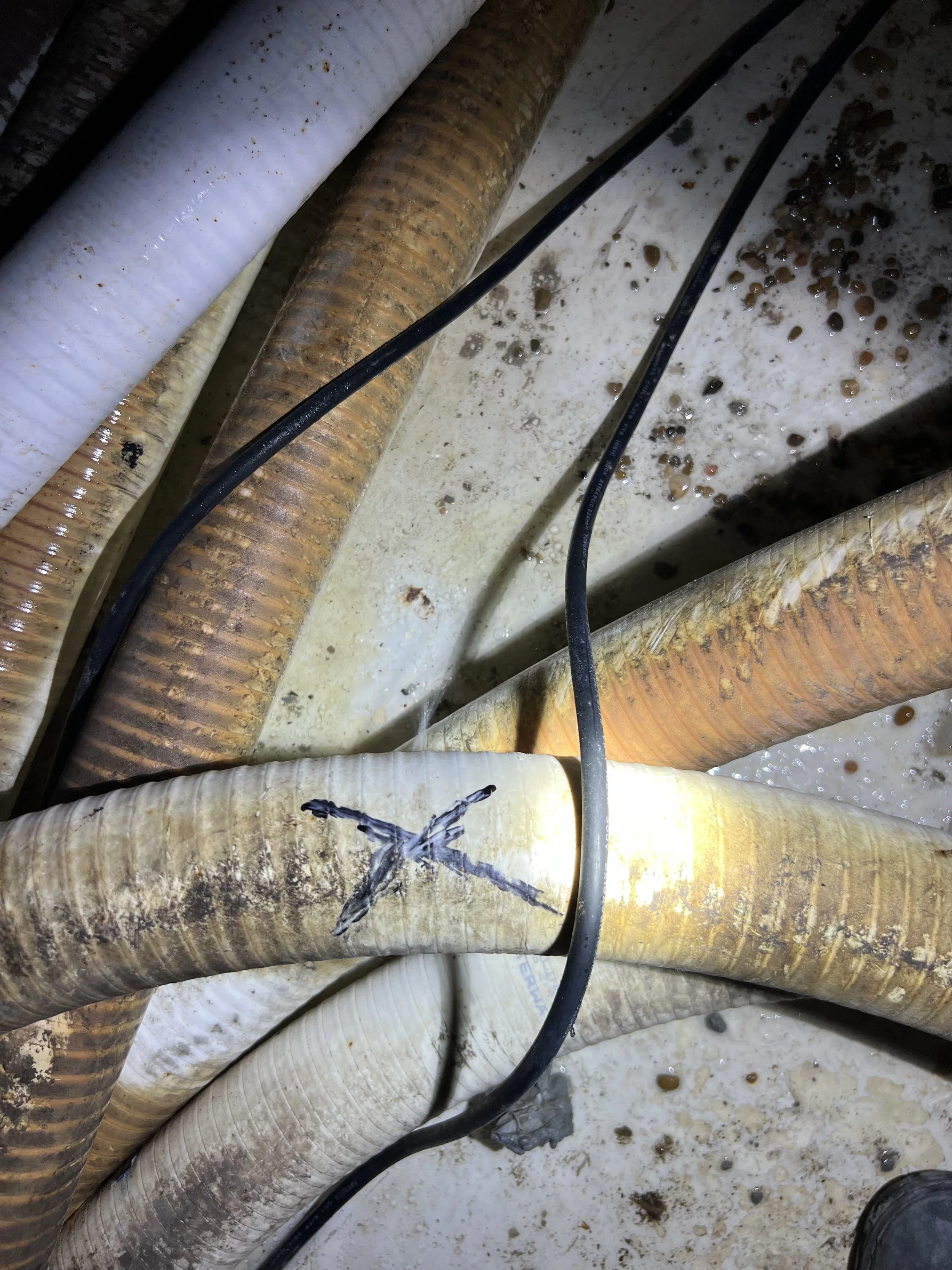 Multiple large pipes or hoses, some white and some rusty brown, lying on a dirty ground with liquid and debris, possibly in a basement or industrial setting.