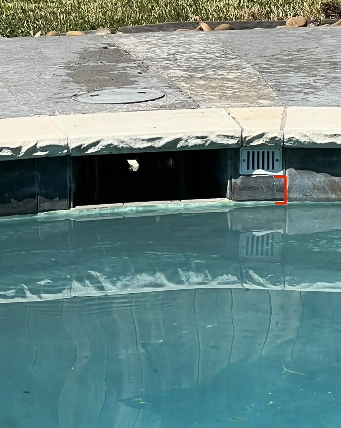 Close-up of a poolside drainage vent with a grate, set against the edge of a swimming pool with a stone deck and some greenery in the background.