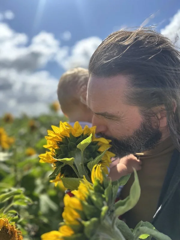 A man with long dark hair and a beard is smelling a sunflower in a sunflower field on a sunny day with a partly cloudy sky.