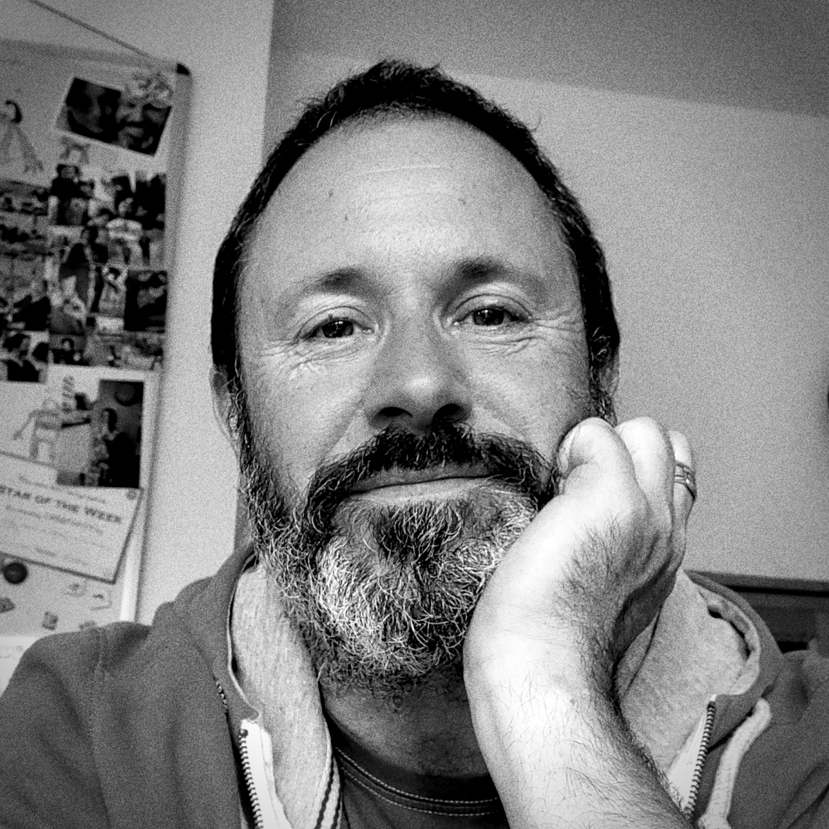 A black-and-white close-up photo of a middle-aged man with a beard, smiling gently with his head resting on his hand, in an indoor setting.