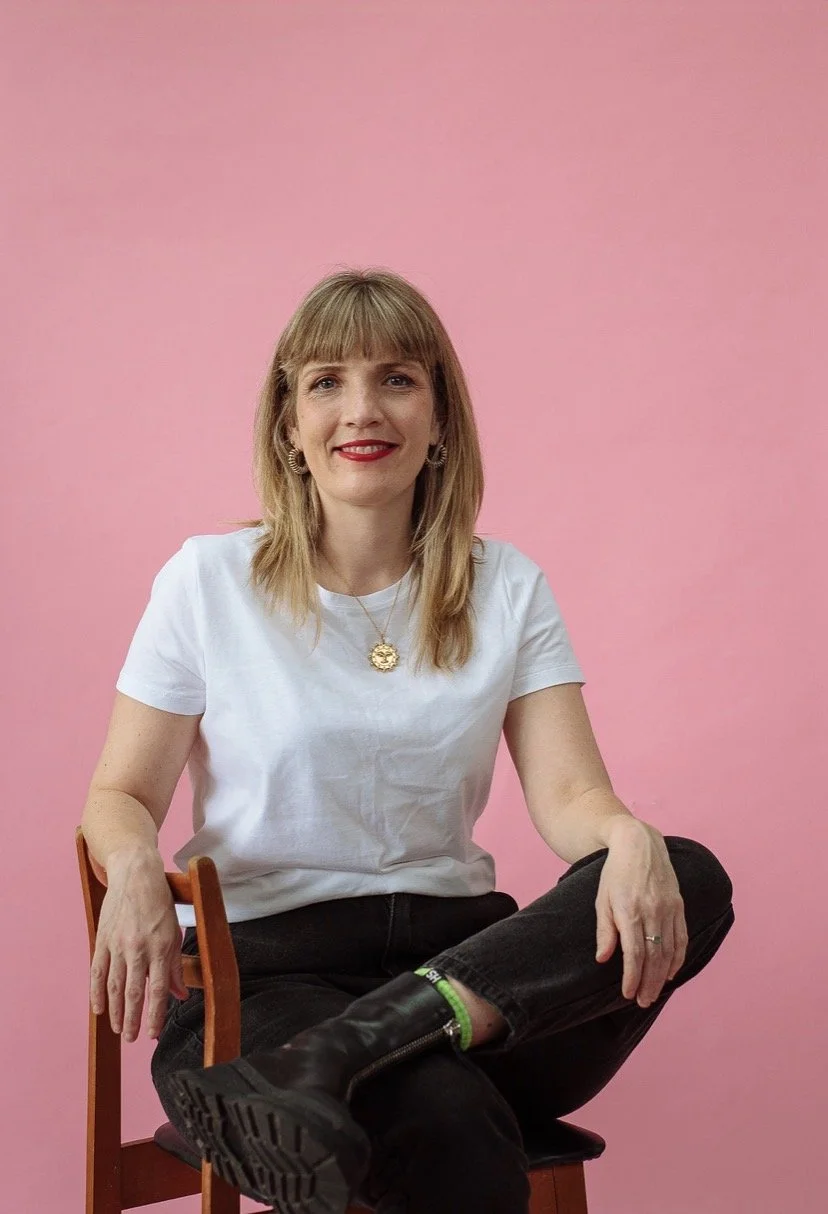 A woman sitting cross-legged on a wooden chair against a pink background, wearing a white t-shirt, black pants, black boots, and gold jewelry, smiling at the camera.