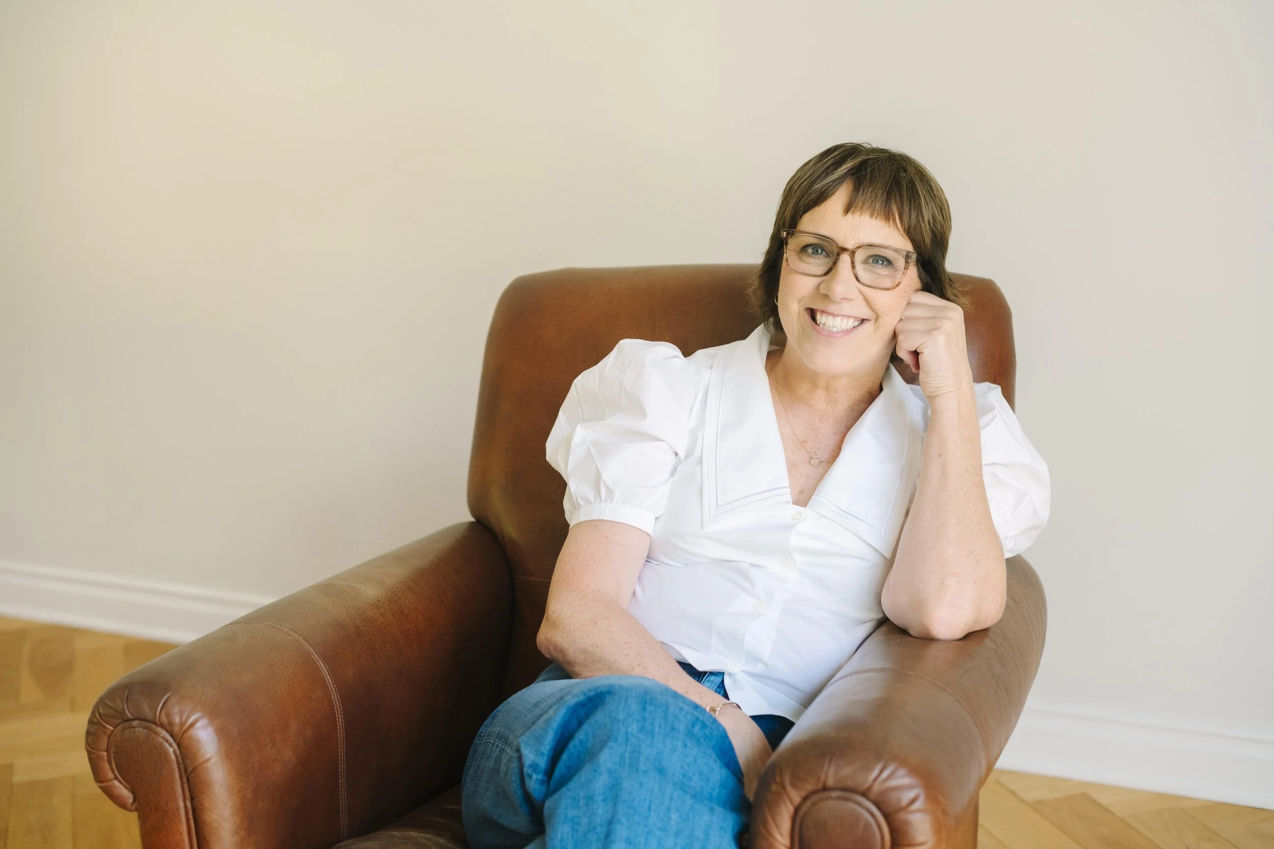 A woman with short brown hair, glasses, and a white shirt sitting on a brown leather couch, smiling at the camera, with her chin resting on her hand.