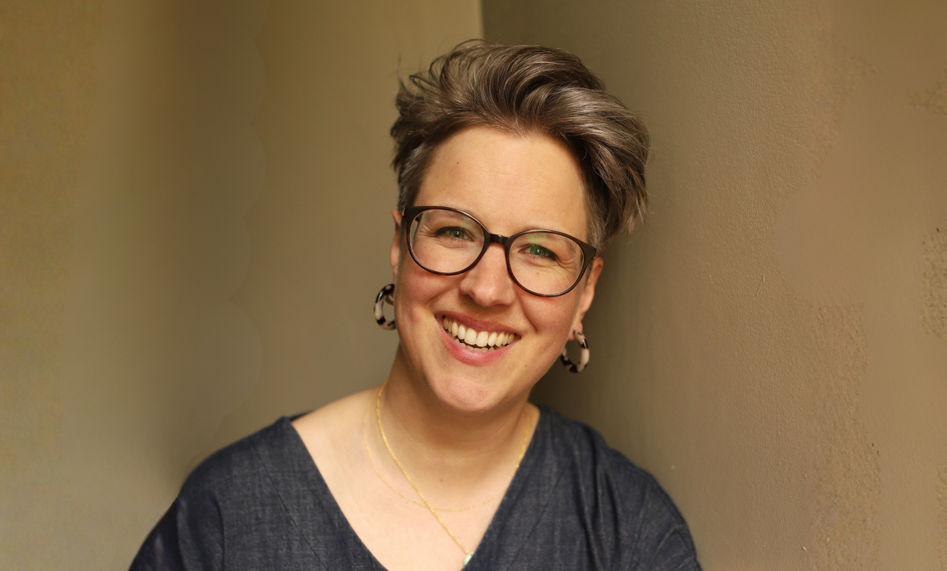 Smiling woman with short gray hair, glasses, hoop earrings, and a dark top, standing against a beige wall.
