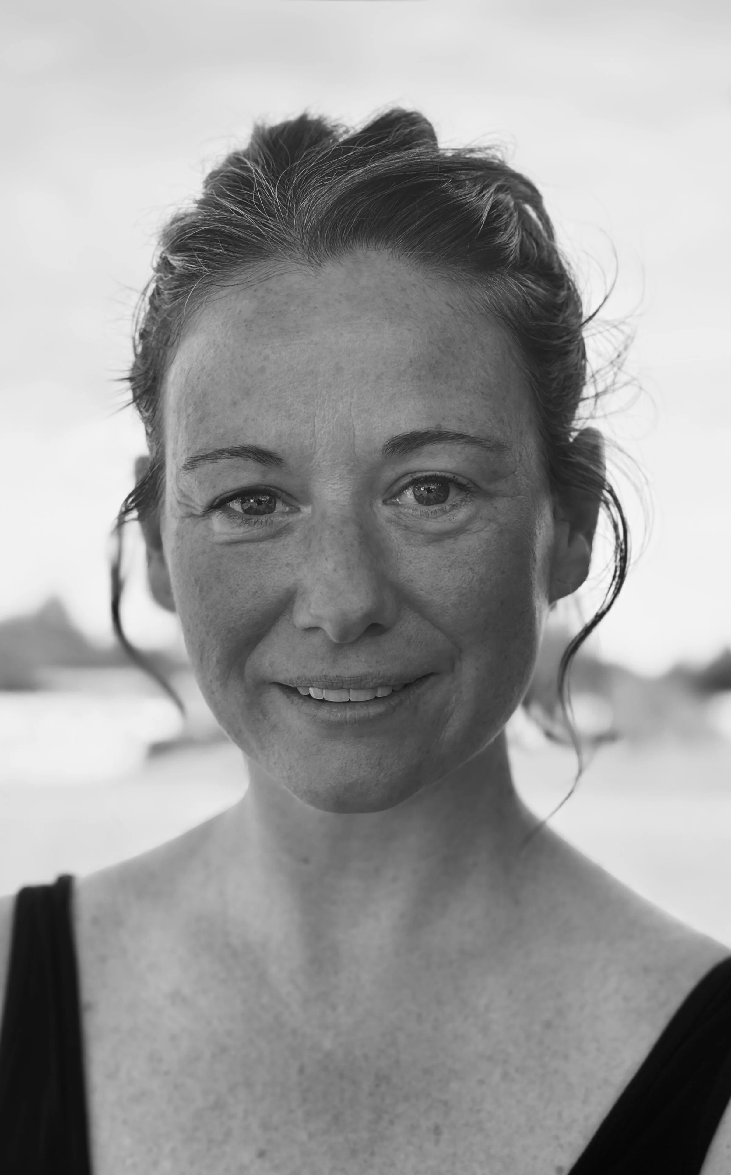 Black and white photo of a smiling woman with exposed shoulders and wearing a dress with thin straps, outdoors with a blurred background.