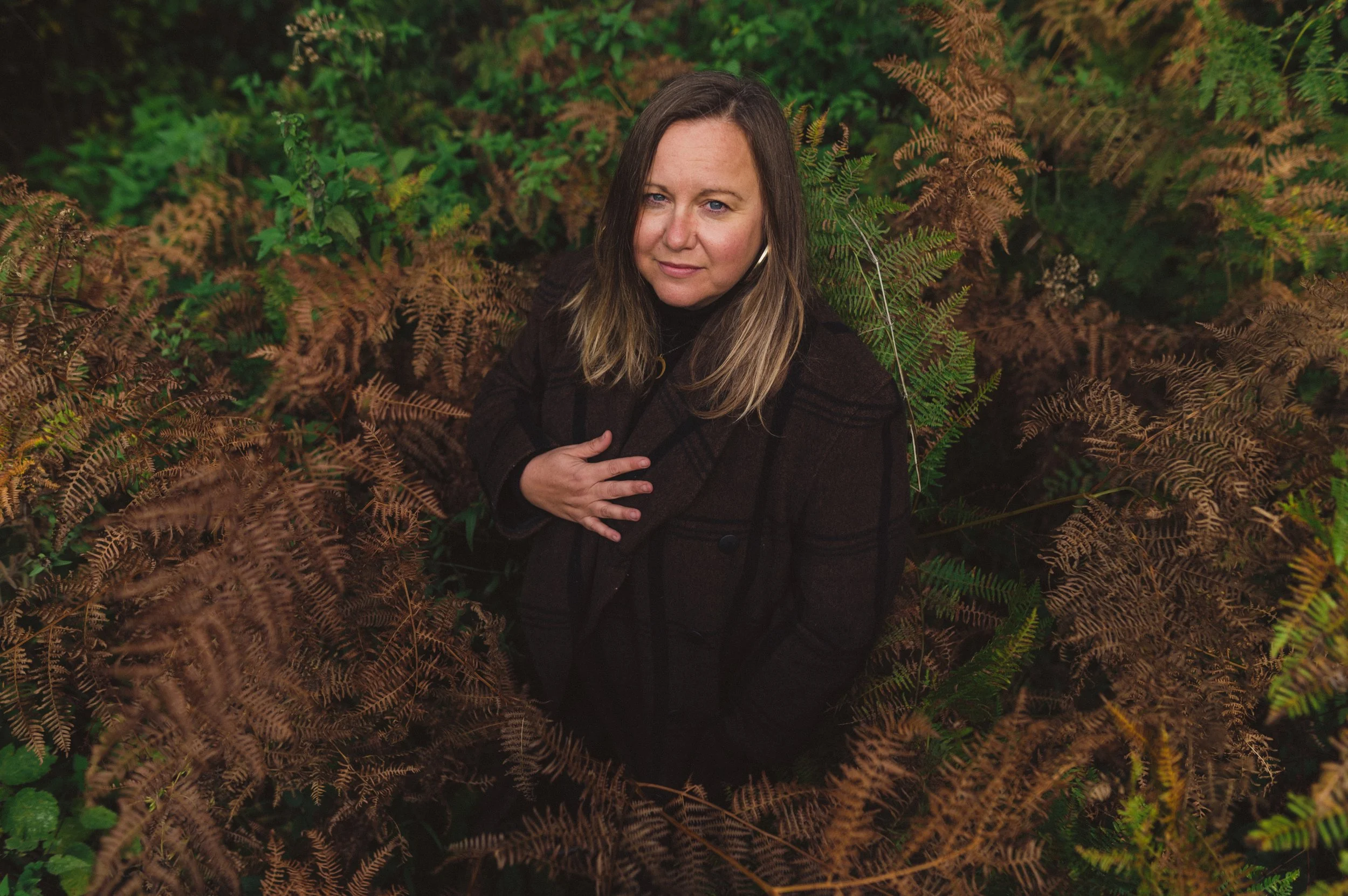 A woman with shoulder-length brown hair is standing amidst lush green and brown fern plants, looking at the camera with a slight smile. She is wearing a black or dark-colored coat.