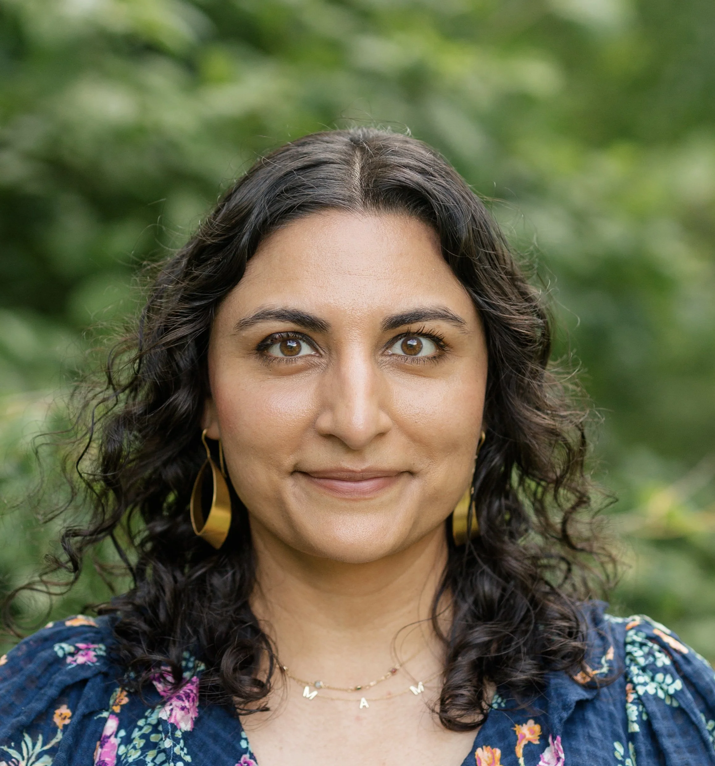 Close-up of a woman with curly dark hair, light brown skin, wearing a blue floral top, gold hoop earrings, and a necklace with the name "MAYA," standing outdoors with a blurred green background.