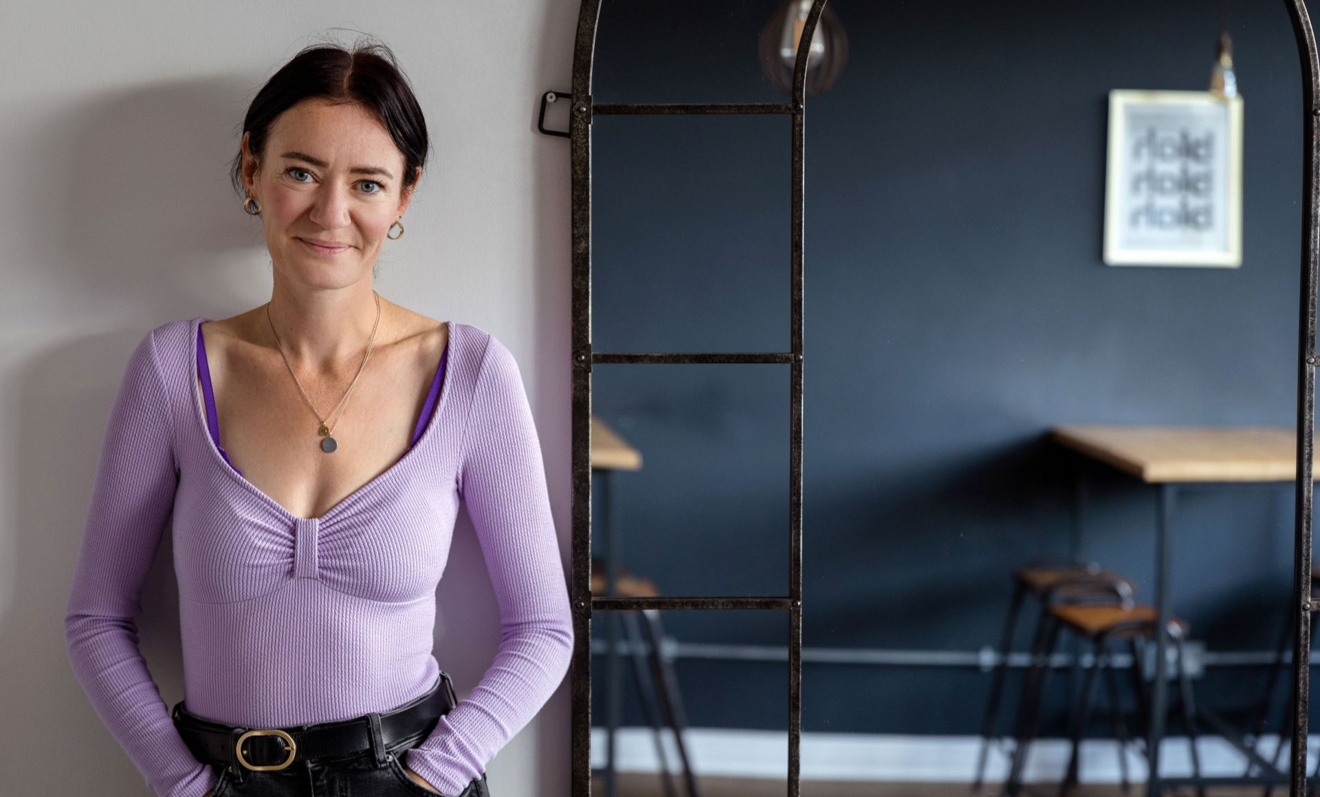 A woman with short dark hair and blue eyes standing indoors, wearing a light purple long-sleeve top and black pants with a belt, smiling slightly at the camera.