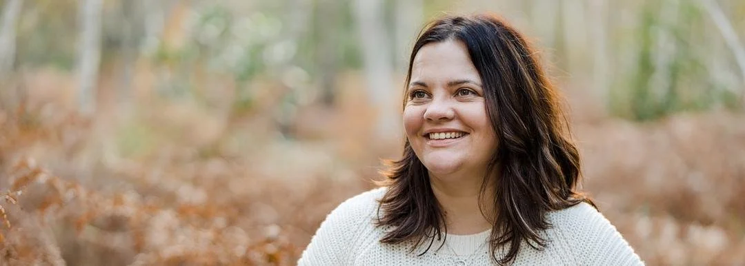 A smiling woman with shoulder-length dark hair standing outdoors in a forested area with fall foliage.