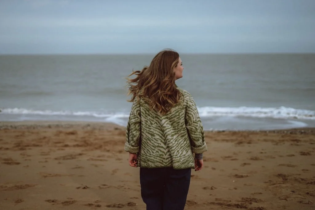 A woman with long, wavy hair stands on a sandy beach facing the ocean, wearing a patterned jacket.