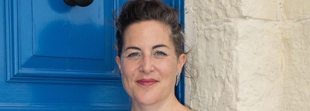 A woman with brown hair and blue eyes is smiling in front of a blue door and beige wall.