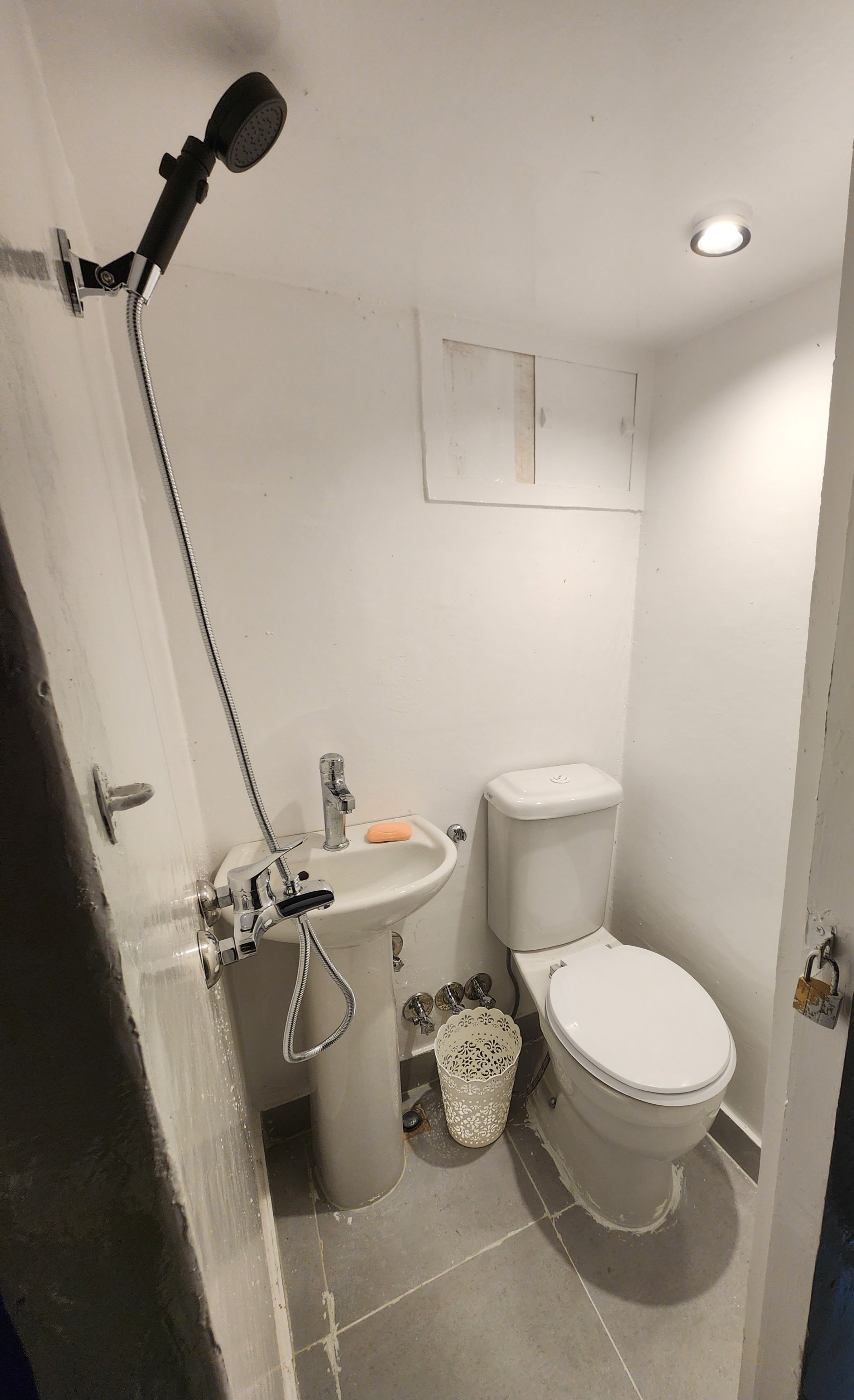 Small bathroom with a toilet, small sink, showerhead on the ceiling, and a wall with a small cabinet. White walls and gray floor tiles.