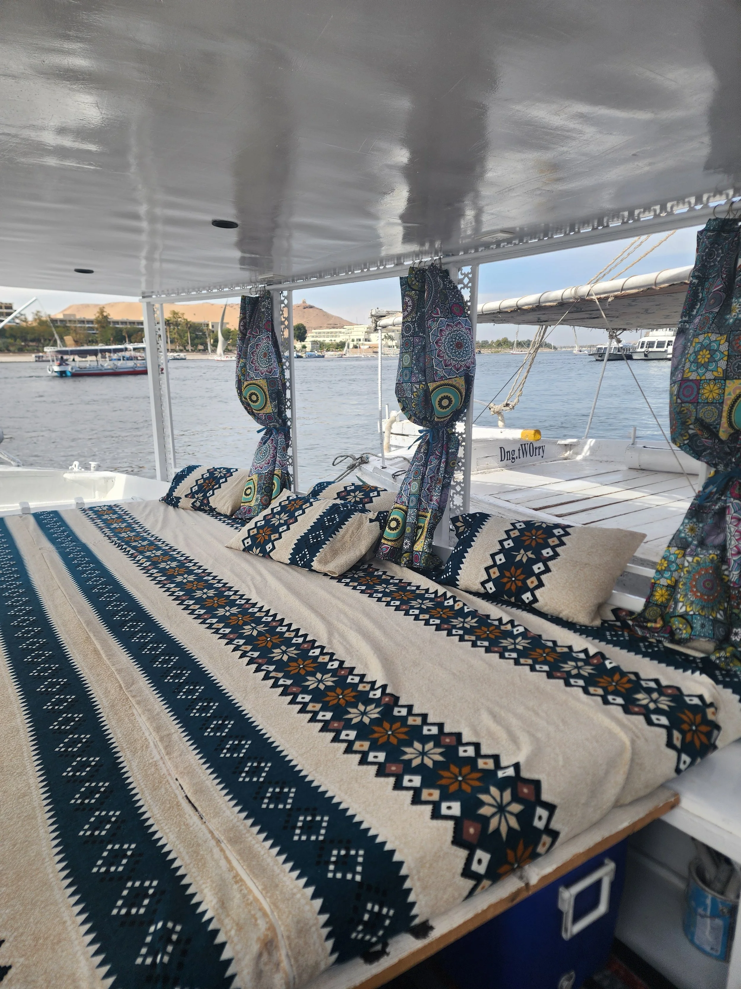 A boat deck with colorful tribal-patterned cushions and curtains, overlooking a river with boats and buildings in the background.