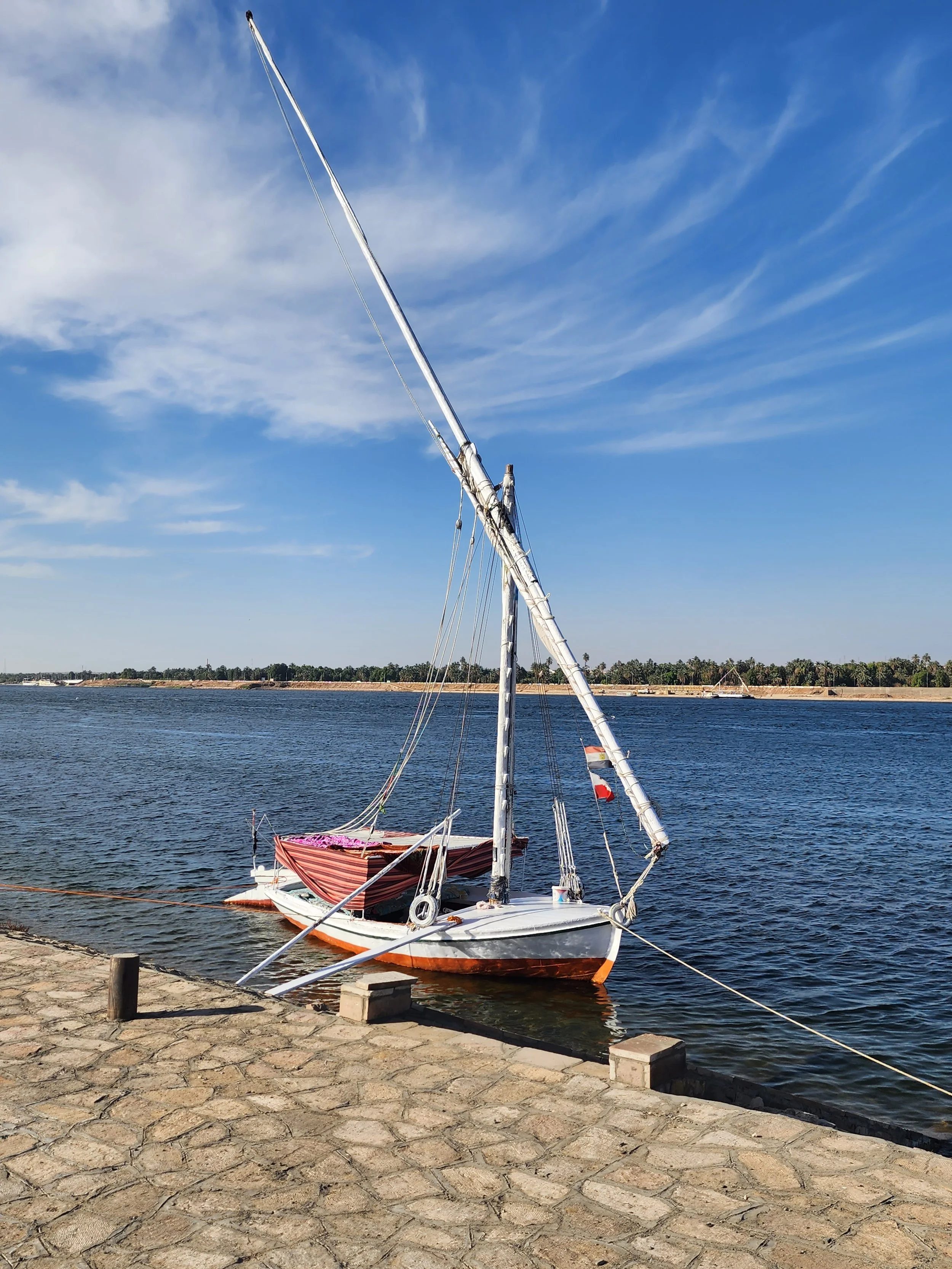 A small sailboat with a partially collapsed sail, docked at the edge of a body of water. The boat has a striped cover on the deck and is tied to a short stone-paved walkway. In the background, there are trees and a distant shoreline under a blue sky with wispy clouds.
