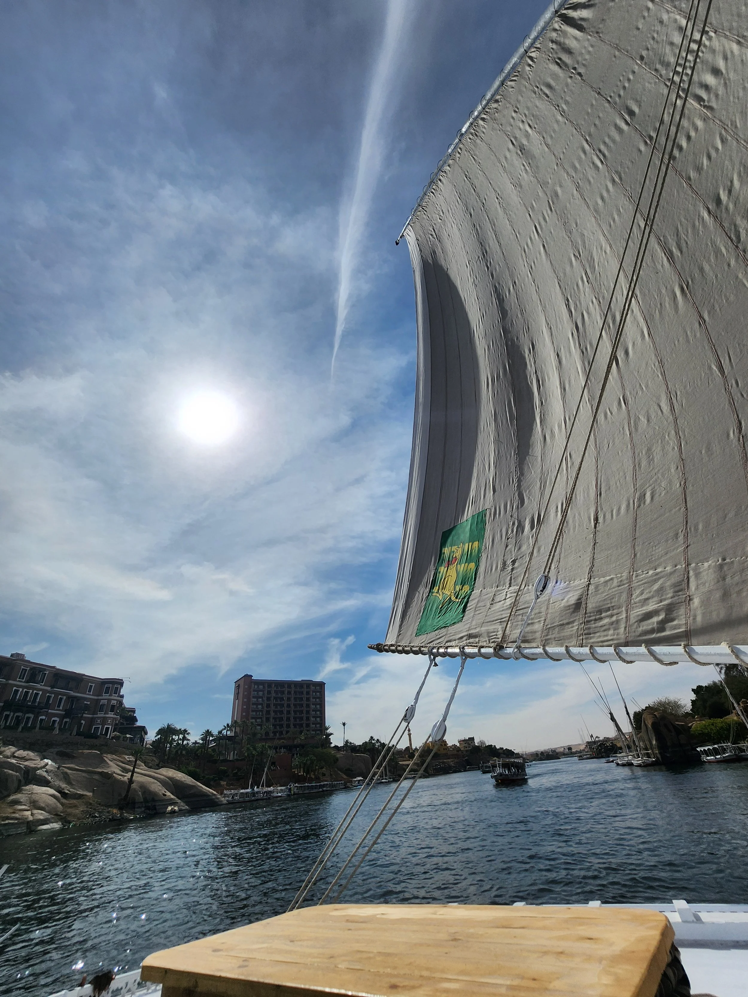 View from a boat looking towards the water with a large sail to the right, buildings along the shoreline, and a partly cloudy sky with the sun shining.