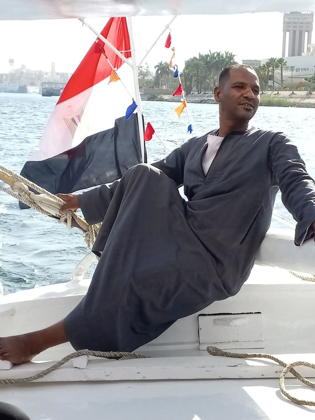A man sitting on a boat with Egyptian flags, wearing a dark robe, with water and city buildings in the background.