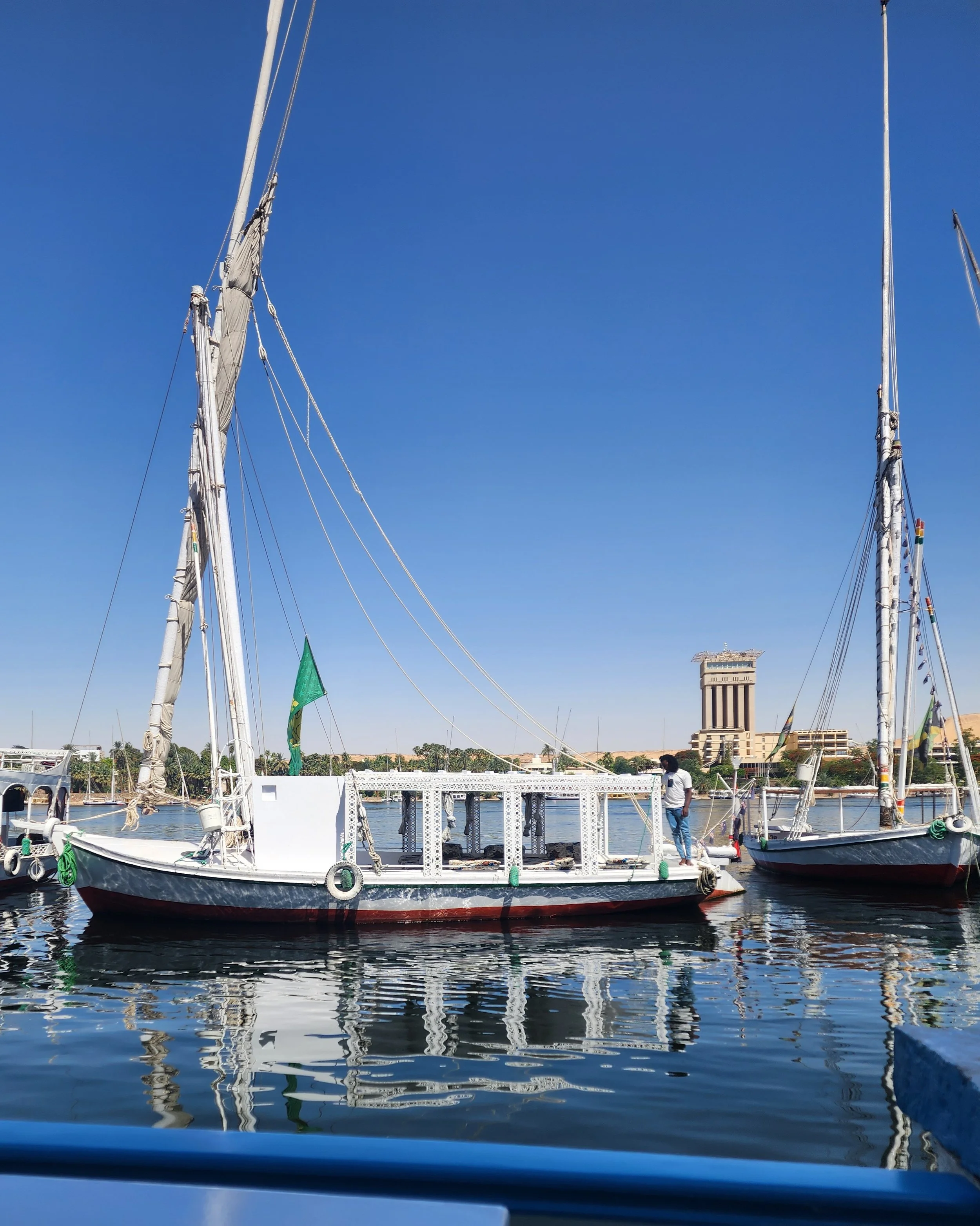 A boat docked on a body of water with two tall masts and sails, and a decorative white structure on the boat. In the background, there is a large building, trees, and a clear blue sky.