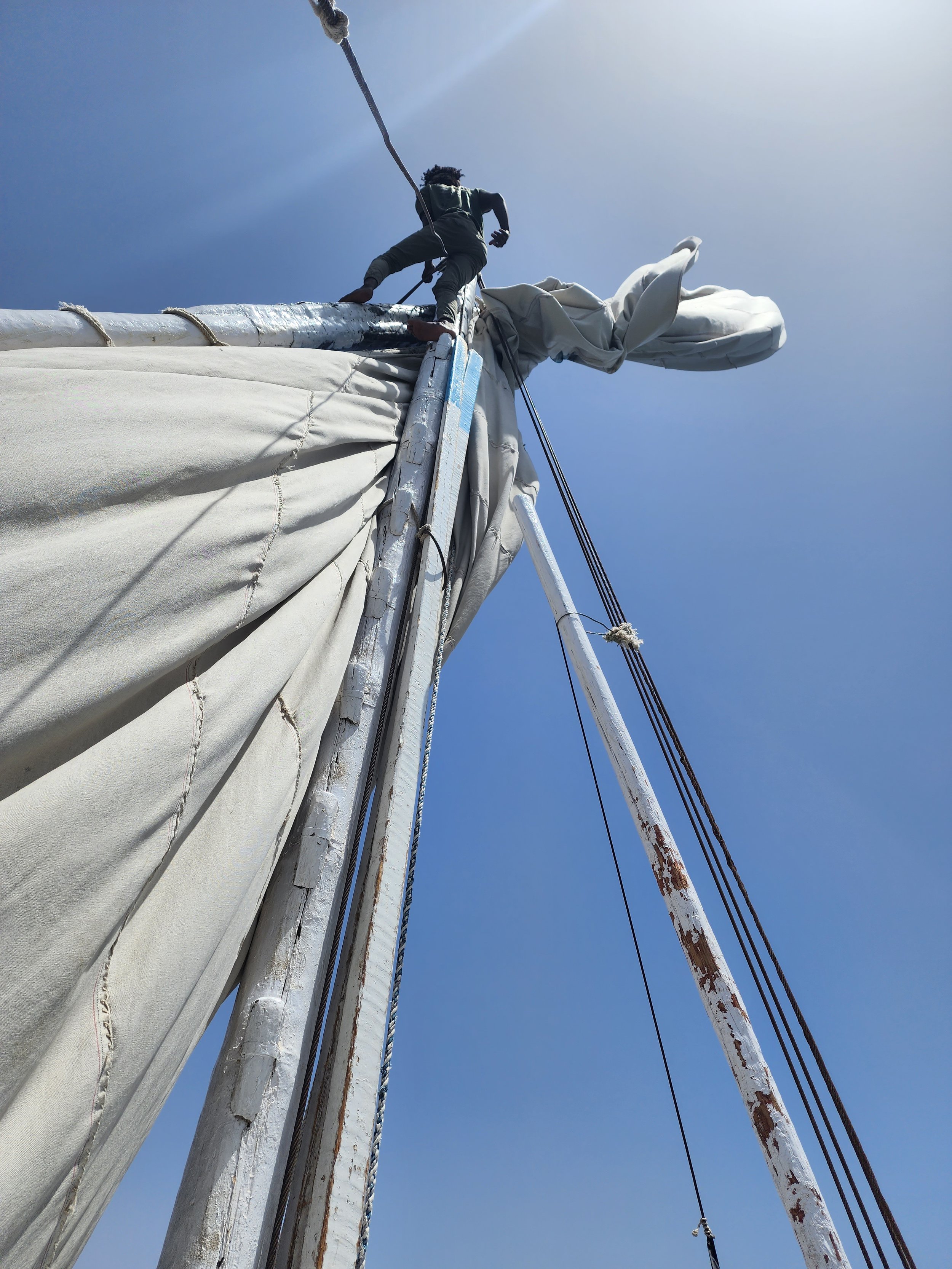 A person is climbing the mast of a sailboat, with the sail and rigging visible against a clear blue sky.