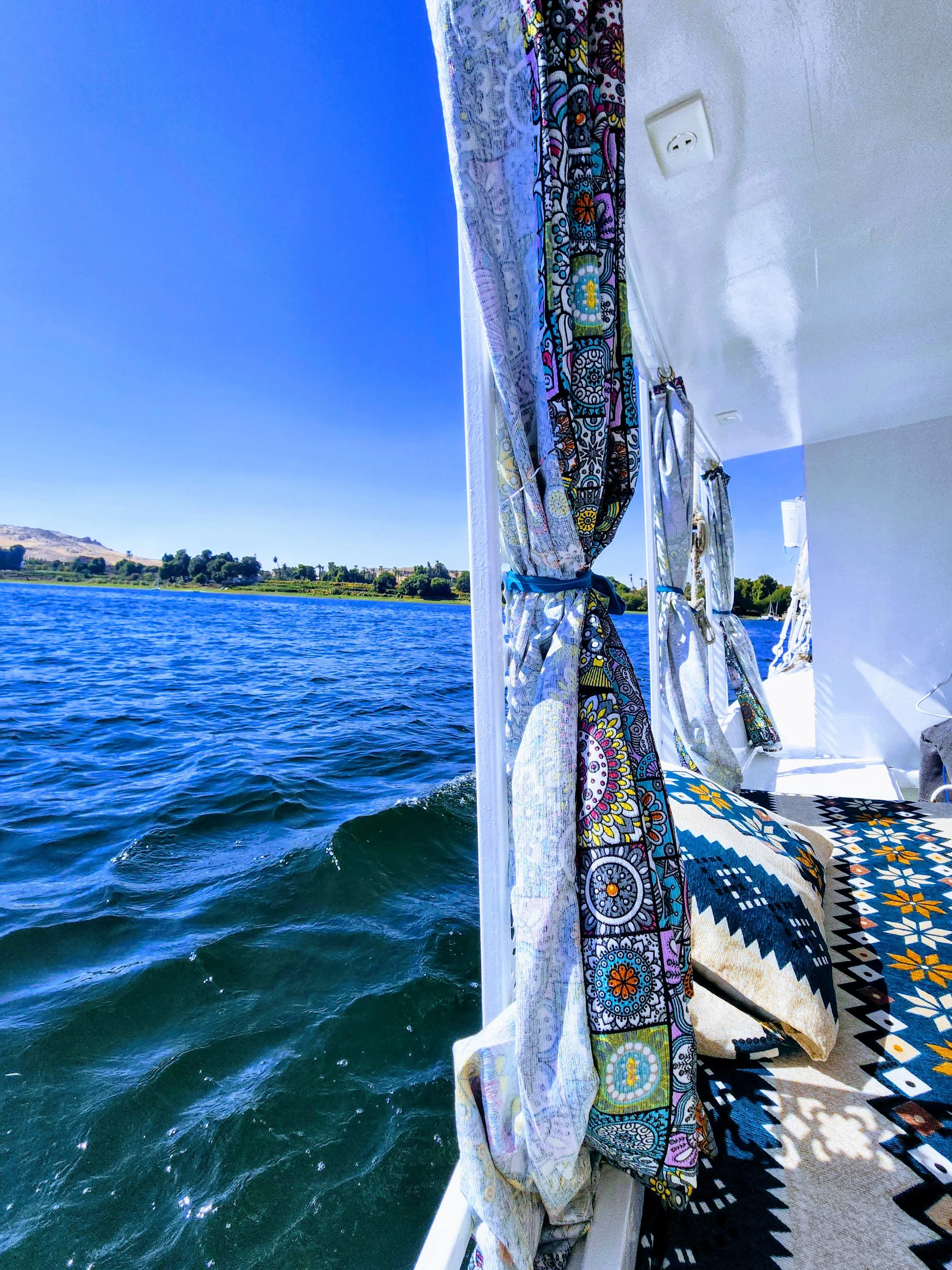 View from a boat showing colorful patterned curtains and a rug on the side, with a body of water and trees in the background under a clear blue sky.