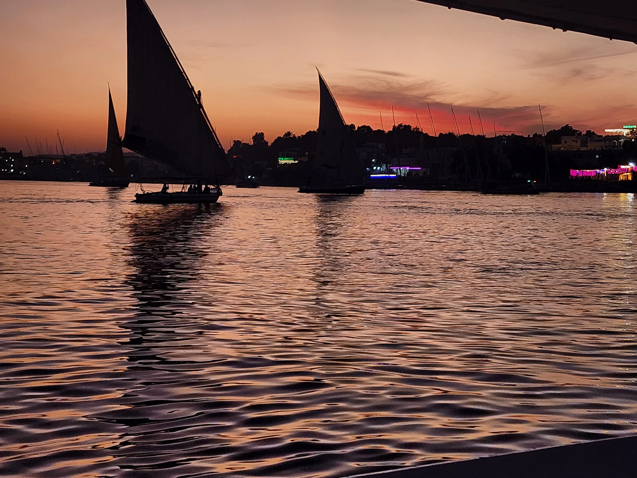 Silhouettes of sailboats on calm water during a sunset with a colorful sky.