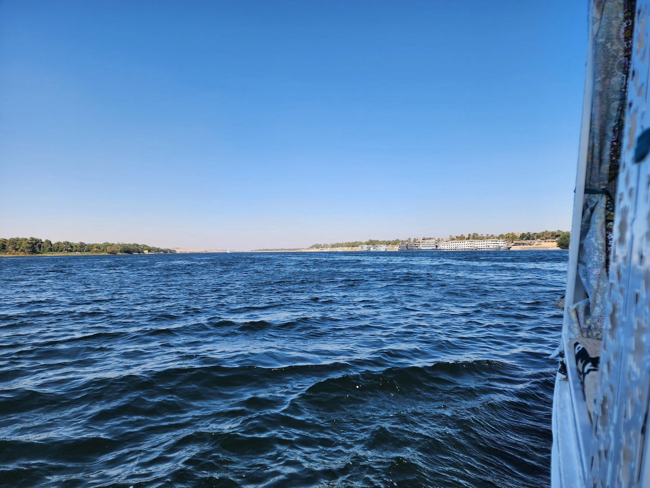 A body of water with gentle ripples under a clear blue sky. On the right, part of a sailboat's side with patterned fabric and a person's foot in a sandal visible.
