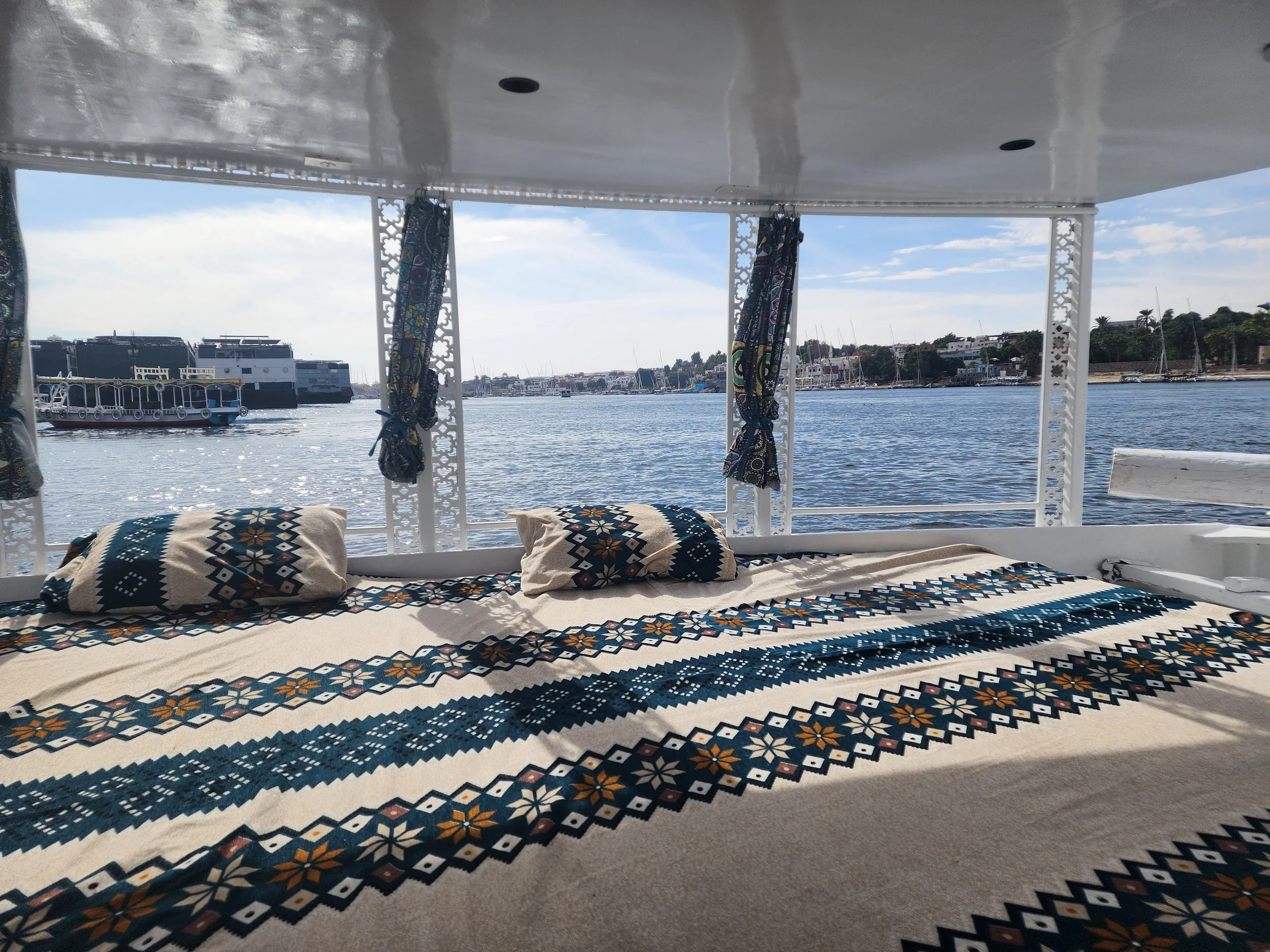 View from inside a boat or yacht, looking out over calm water with docked sailboats and buildings on the shoreline in the distance. The boat's interior has patterned pillows and curtains.