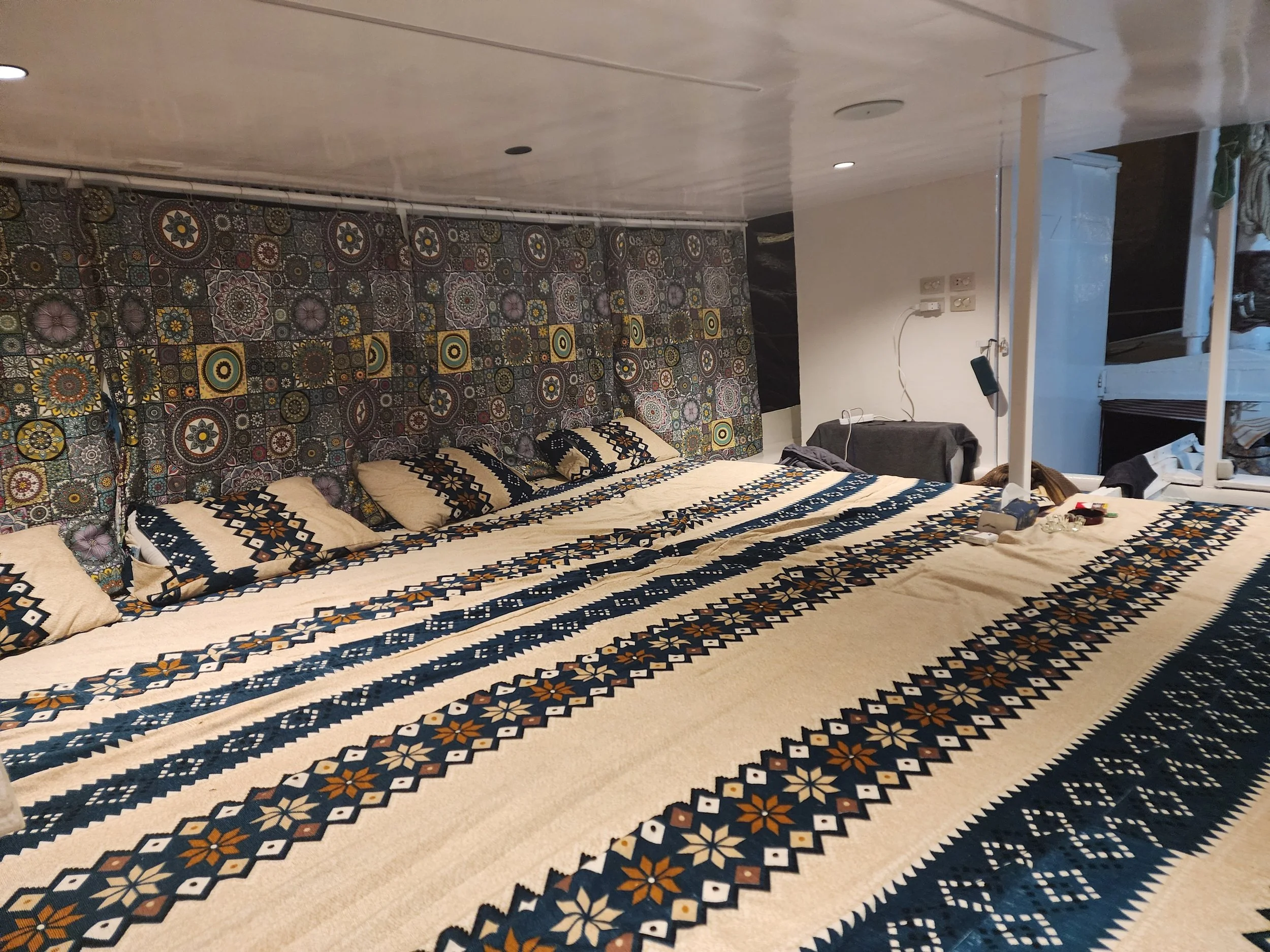 A bedroom with a bed covered in a patterned bedspread and matching pillows, a wall decorated with colorful tiles, a power outlet on the wall, and various items on a bedside table.
