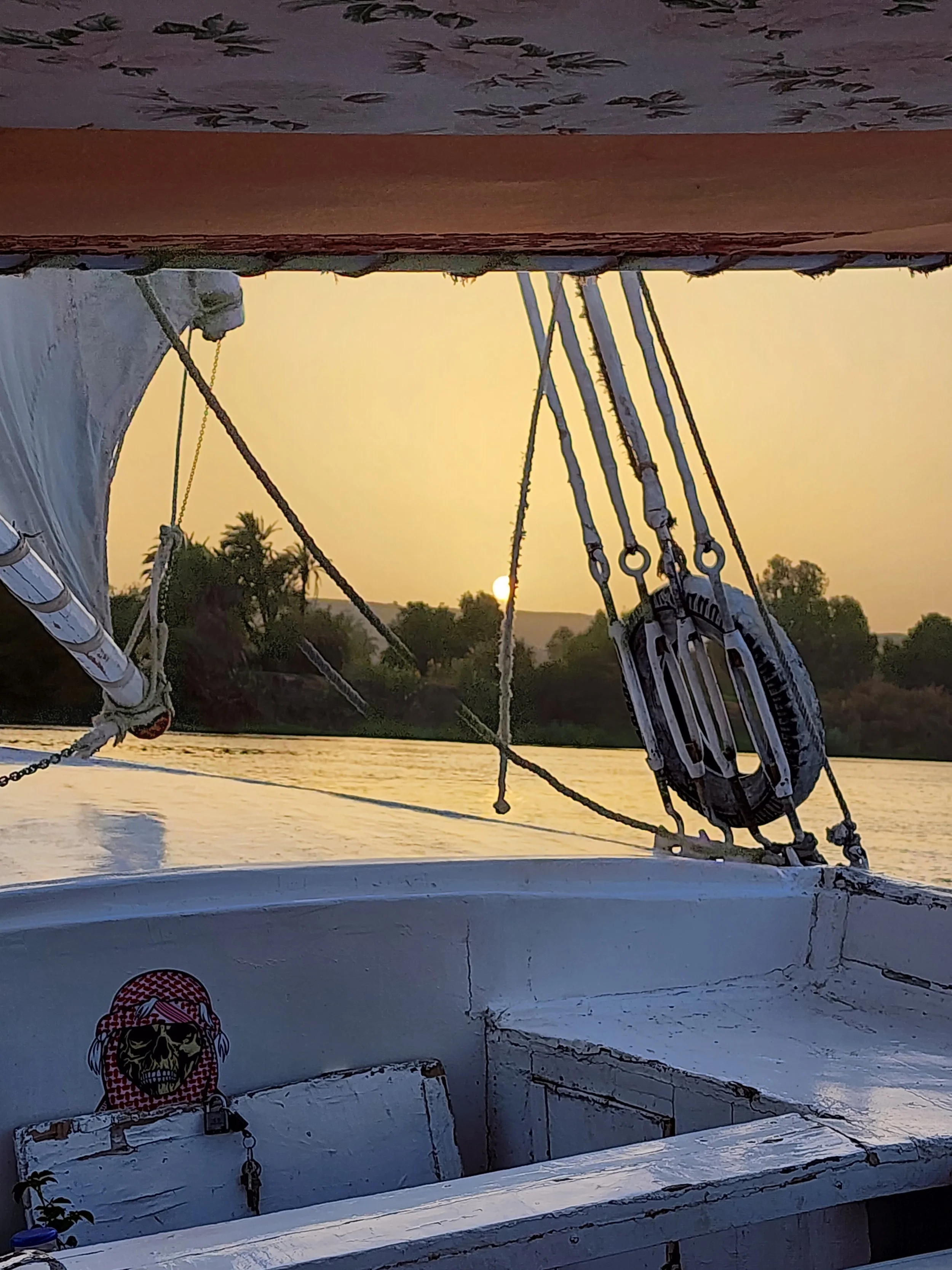 A boat's deck and mast silhouetted against a setting sun over a river, with trees in the distance and a sticker of a skull with sunglasses and a red bandana on the boat's panel.