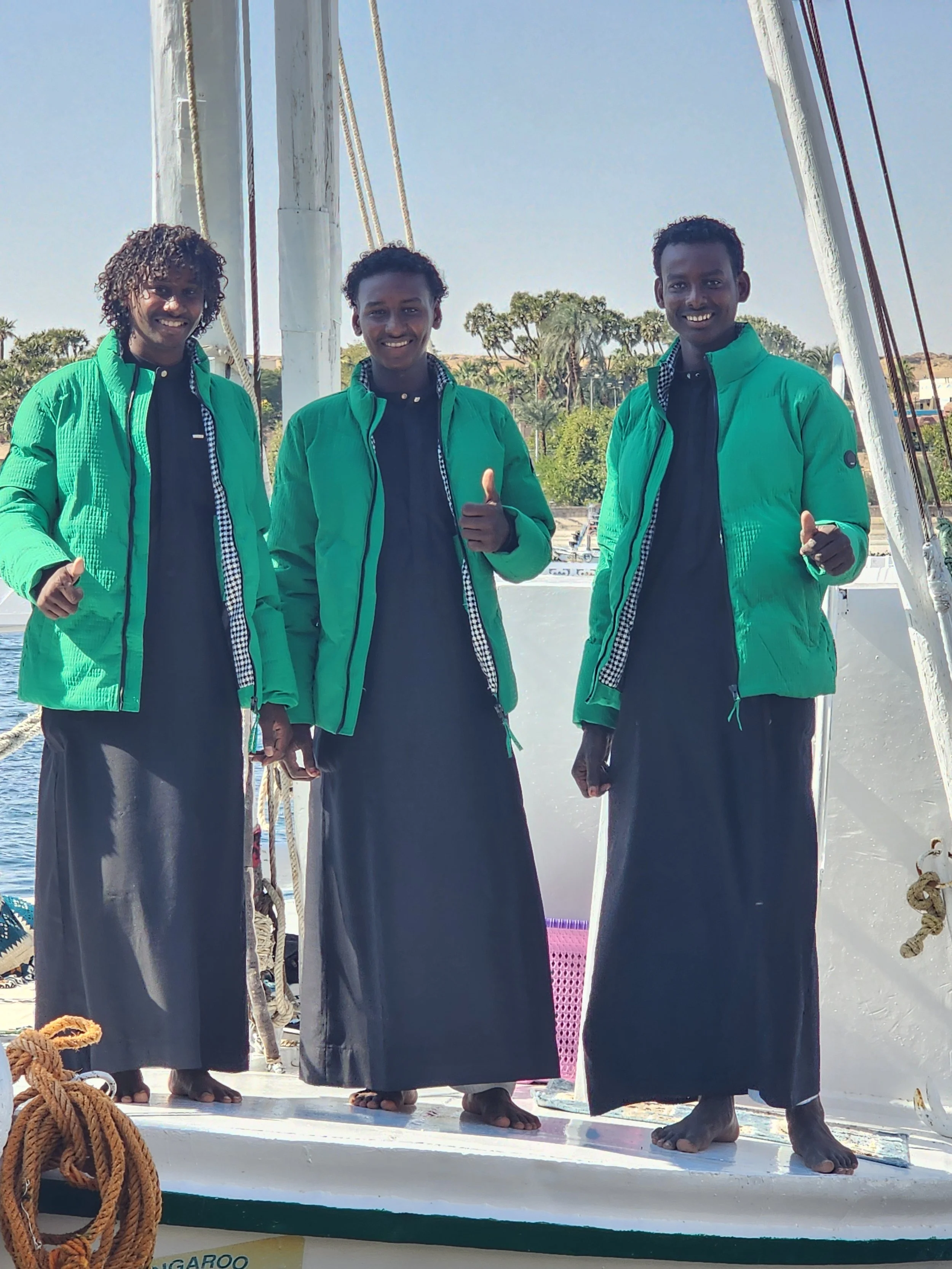Three young men standing on a boat, smiling and giving thumbs-up gestures, dressed in matching green jackets and black robes.