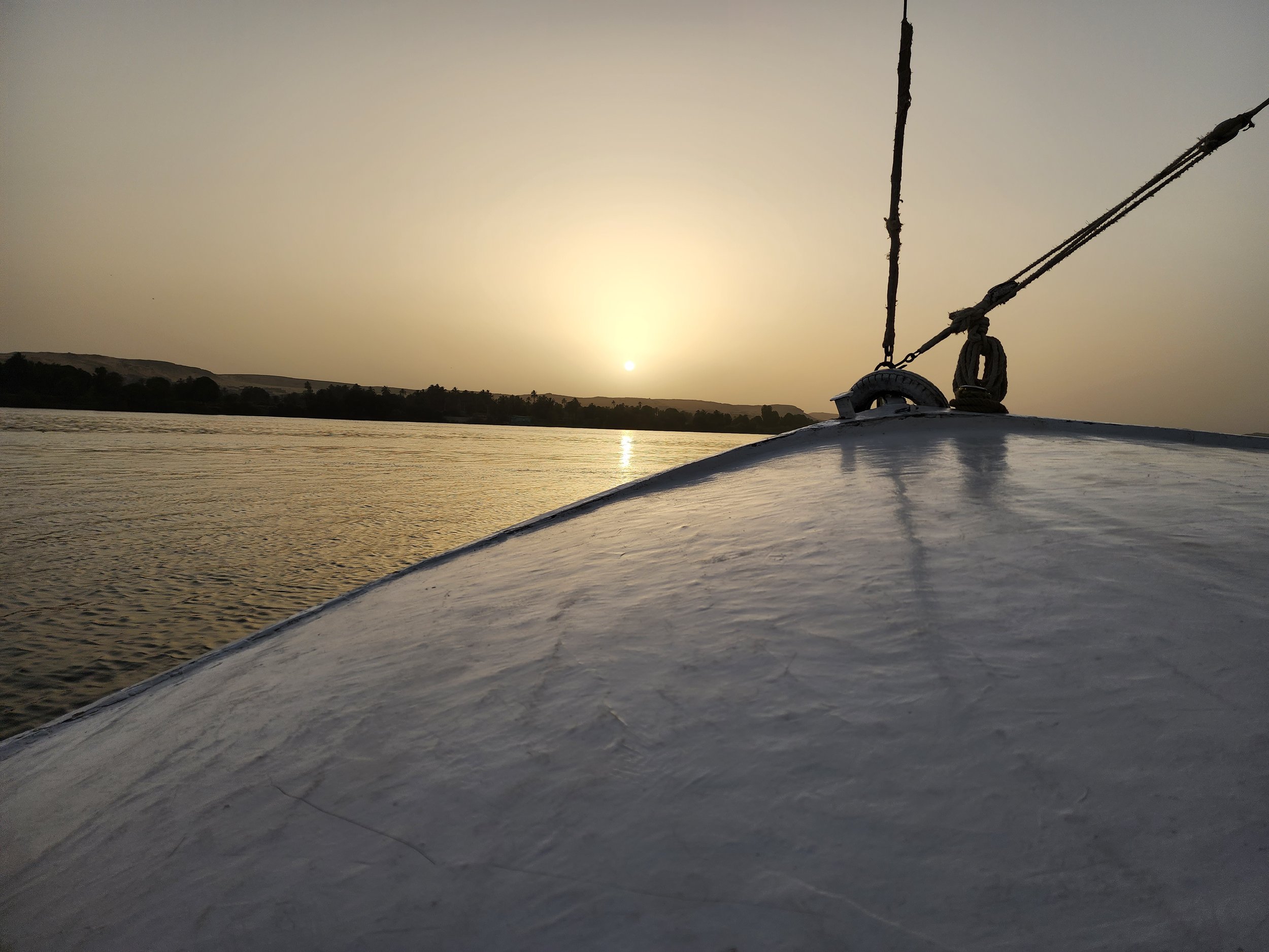 Sunset over a body of water, viewed from the deck of a boat with a metal railing and ropes.