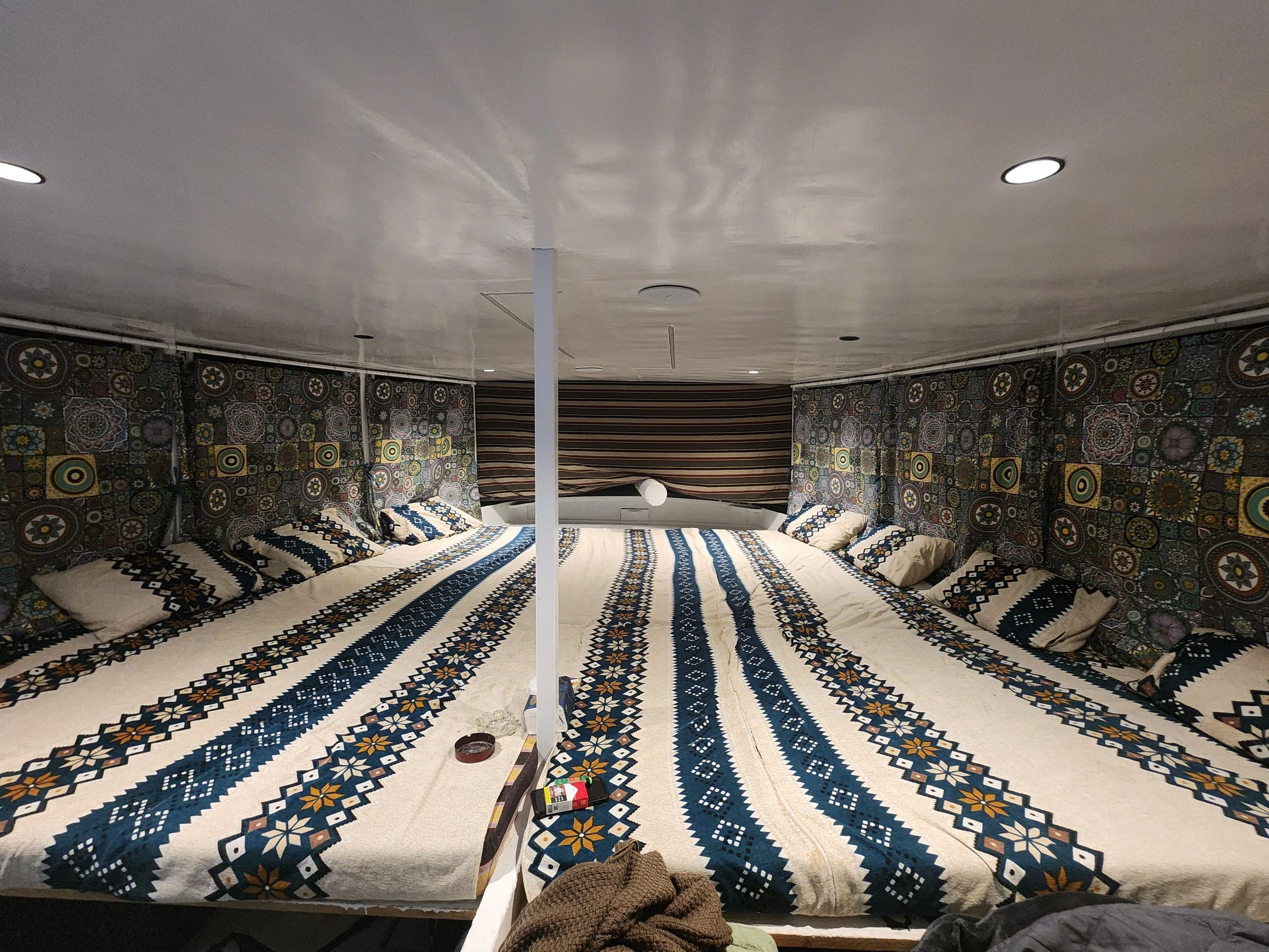 The image shows the interior of a room with a large, patterned rug covering the entire floor. The ceiling is plain with several recessed lights, and the walls are covered with colorful, geometric, and floral patterned fabric or wallpaper. There are some scattered items on the floor, including a red lighter, a box, a pair of shoes, and a pair of socks. A white pole or support runs through the middle of the room, and parts of some furniture or objects are partially visible.