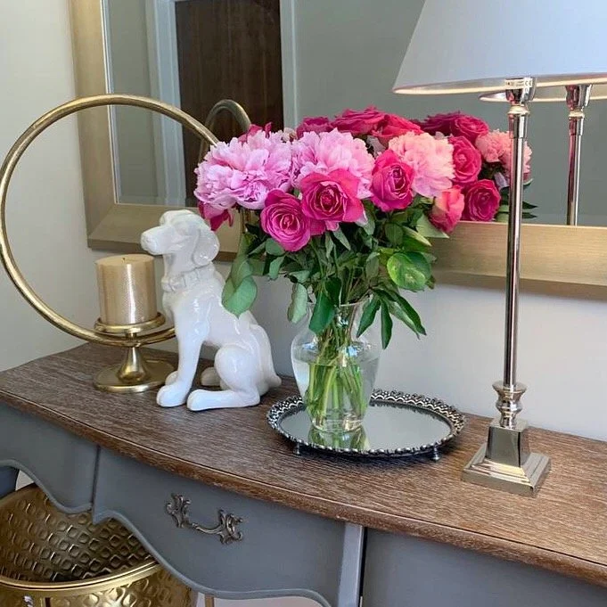 Decorative table with a vase of pink and magenta flowers, a white ceramic dog figurine, a gold candle holder, and a silver tray, with a mirror and a lamp behind.