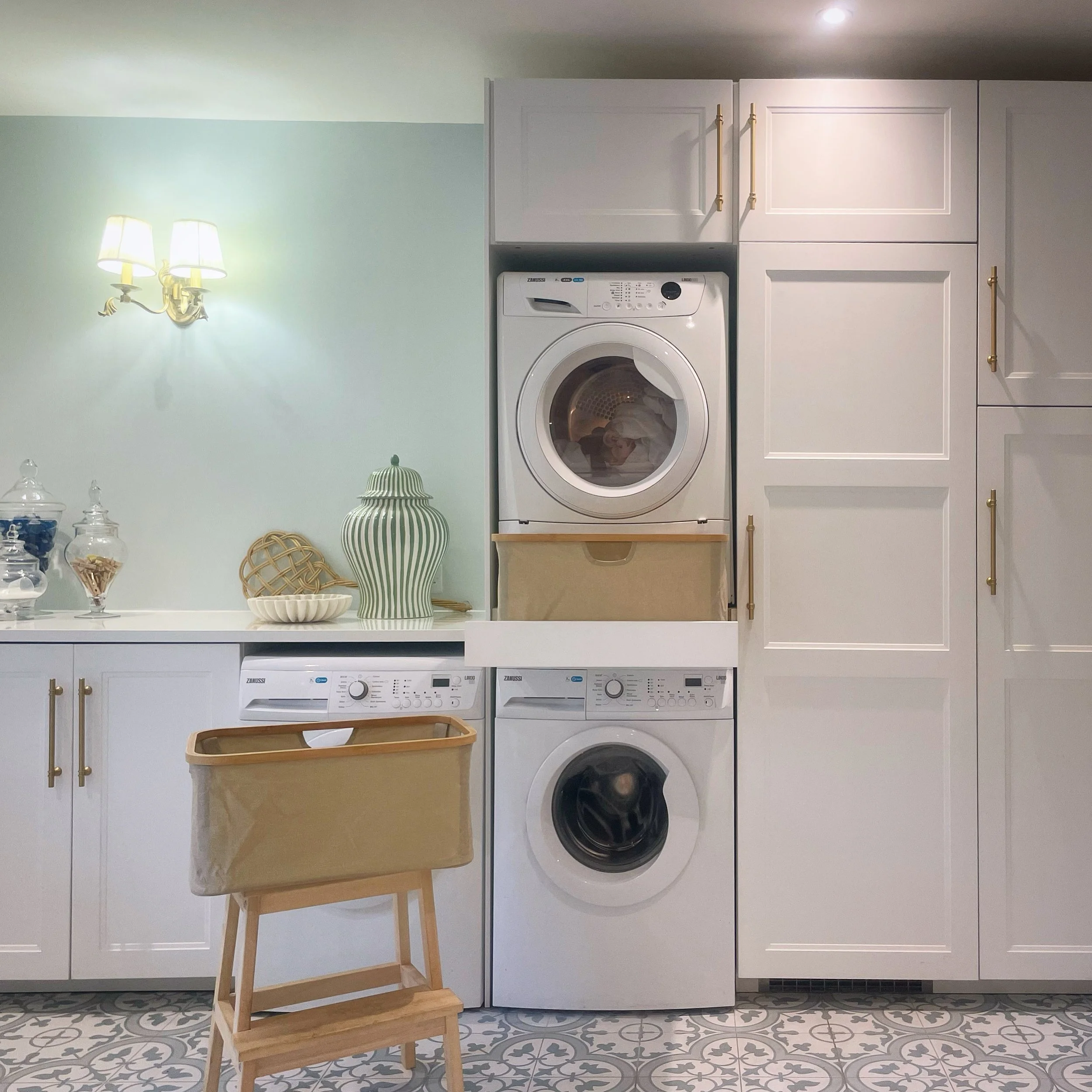 Laundry room with a white washing machine and matching white dryer stacked, white cabinets with gold handles, a small wooden laundry basket, and decorative items, including a striped vase and glass jars, on a patterned tile floor, and a light green wall with a wall-mounted lamp.