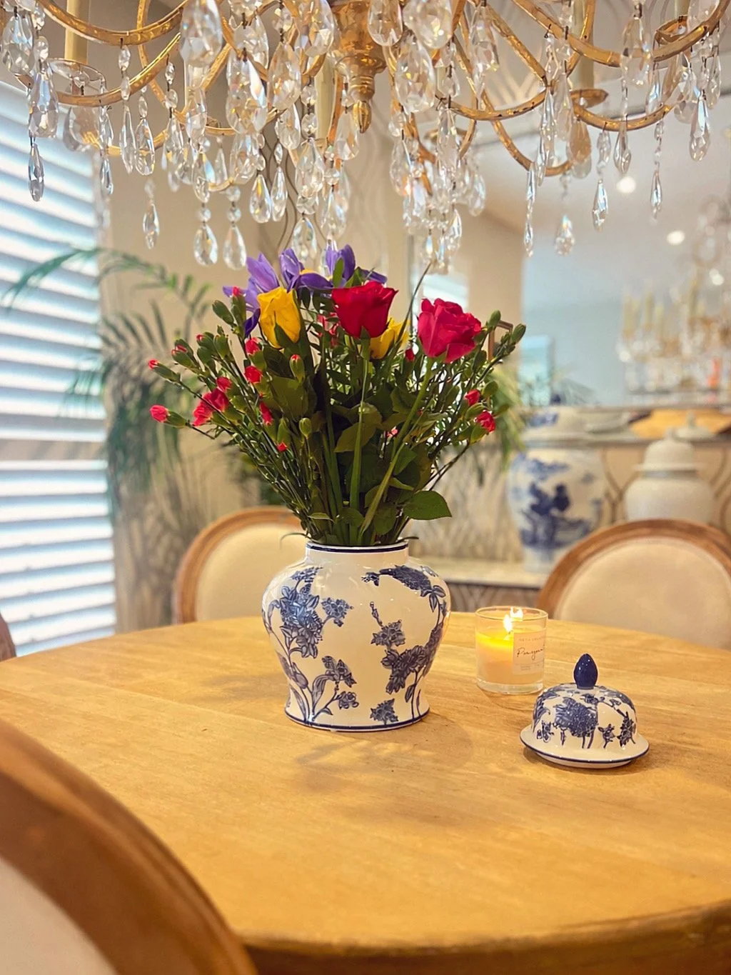 A dining table with a blue and white floral vase holding a colourful bouquet of flowers, and a lit candle, surrounded by elegant chairs and a chandelier with crystal accents.