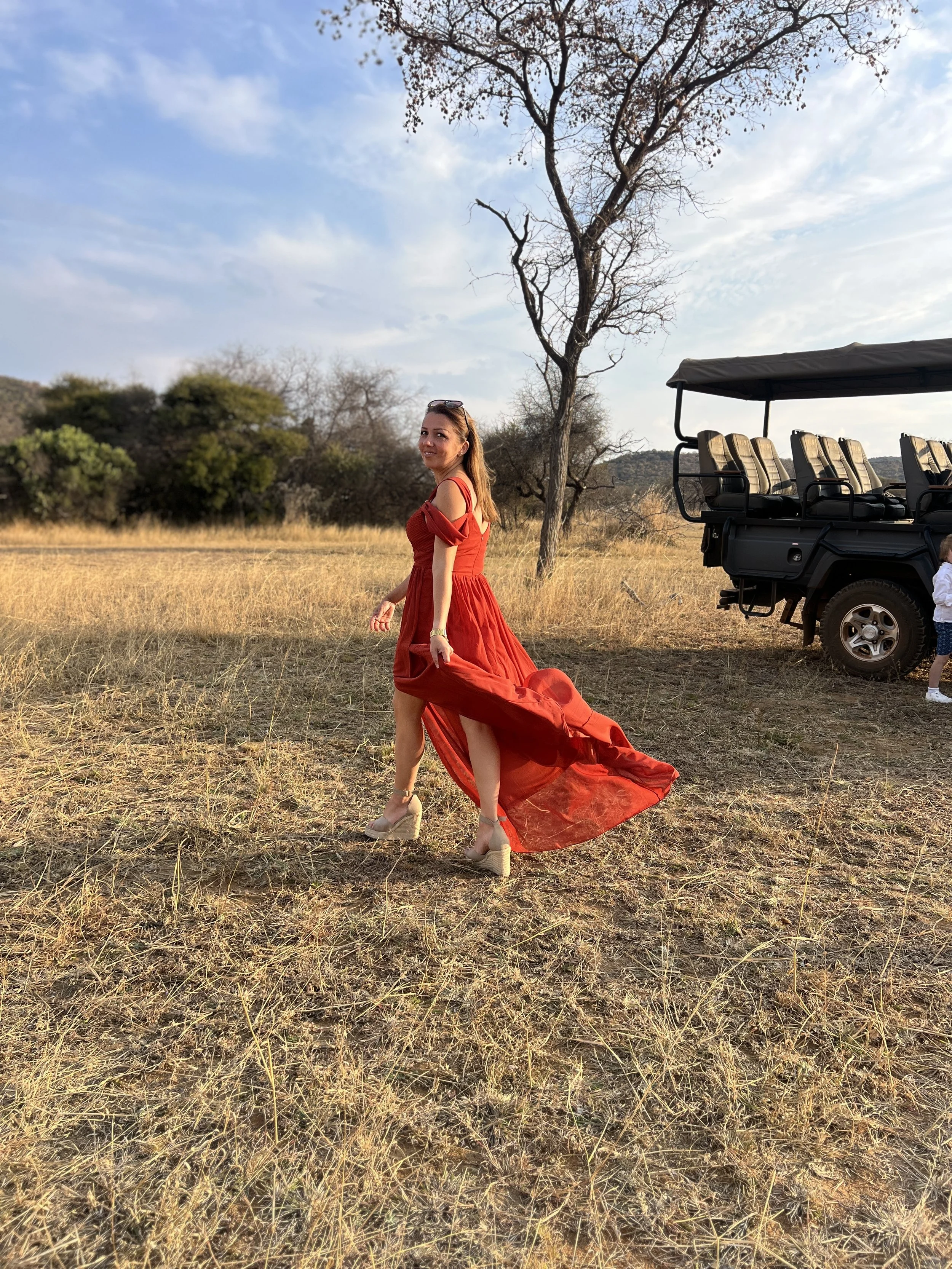 A woman in an orange-red dress with a long train, beige high heels, and sunglasses on her head, standing in a dry grassy field with a leafless tree, and a safari truck. The sky is partly cloudy.