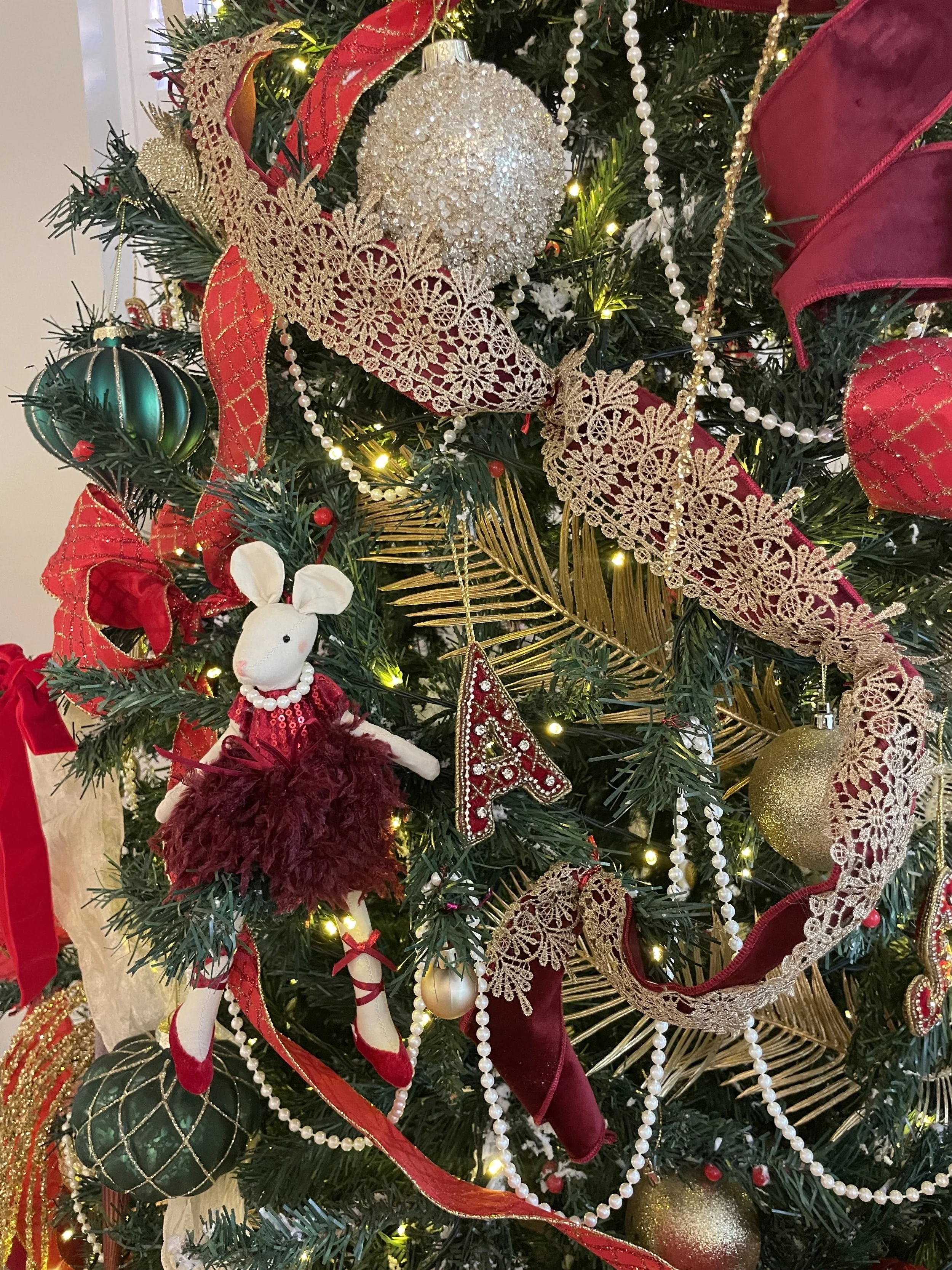 Close-up of a decorated Christmas tree with ornaments including a sparkly white ball, gold and green baubles, a plush mouse wearing a red dress, ribbon, and strings of pearls, illuminated by small warm lights.