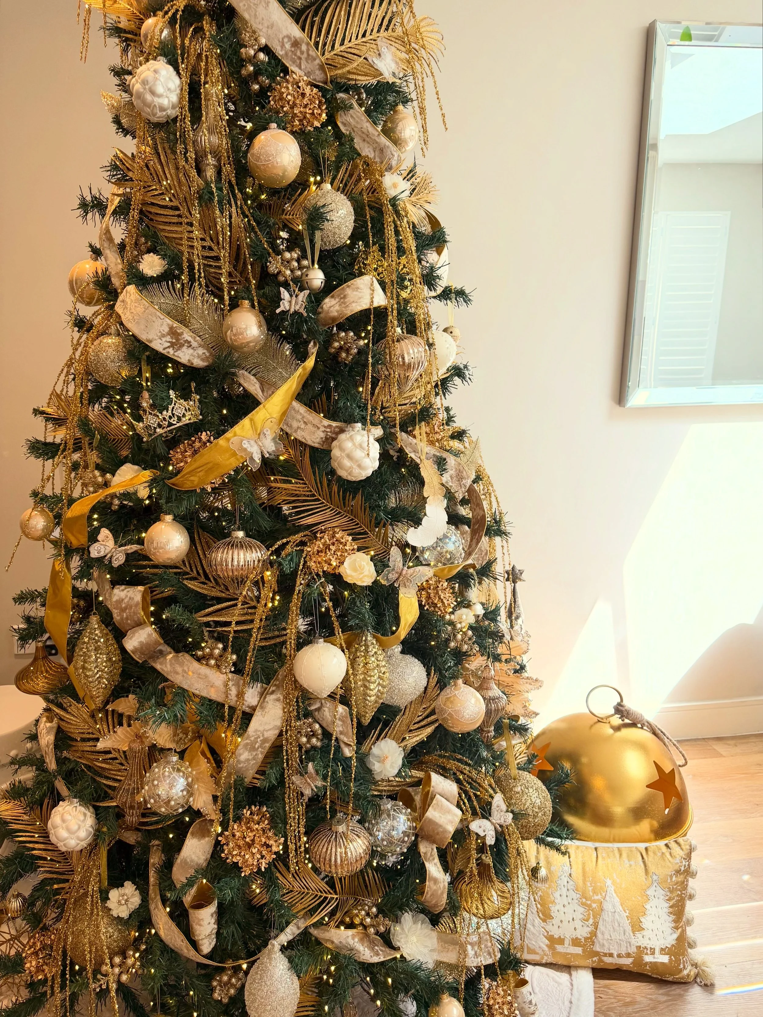 Decorated Christmas tree with gold and white ornaments, ribbons, and lights, placed near a window with a gold and white pillow on the floor.