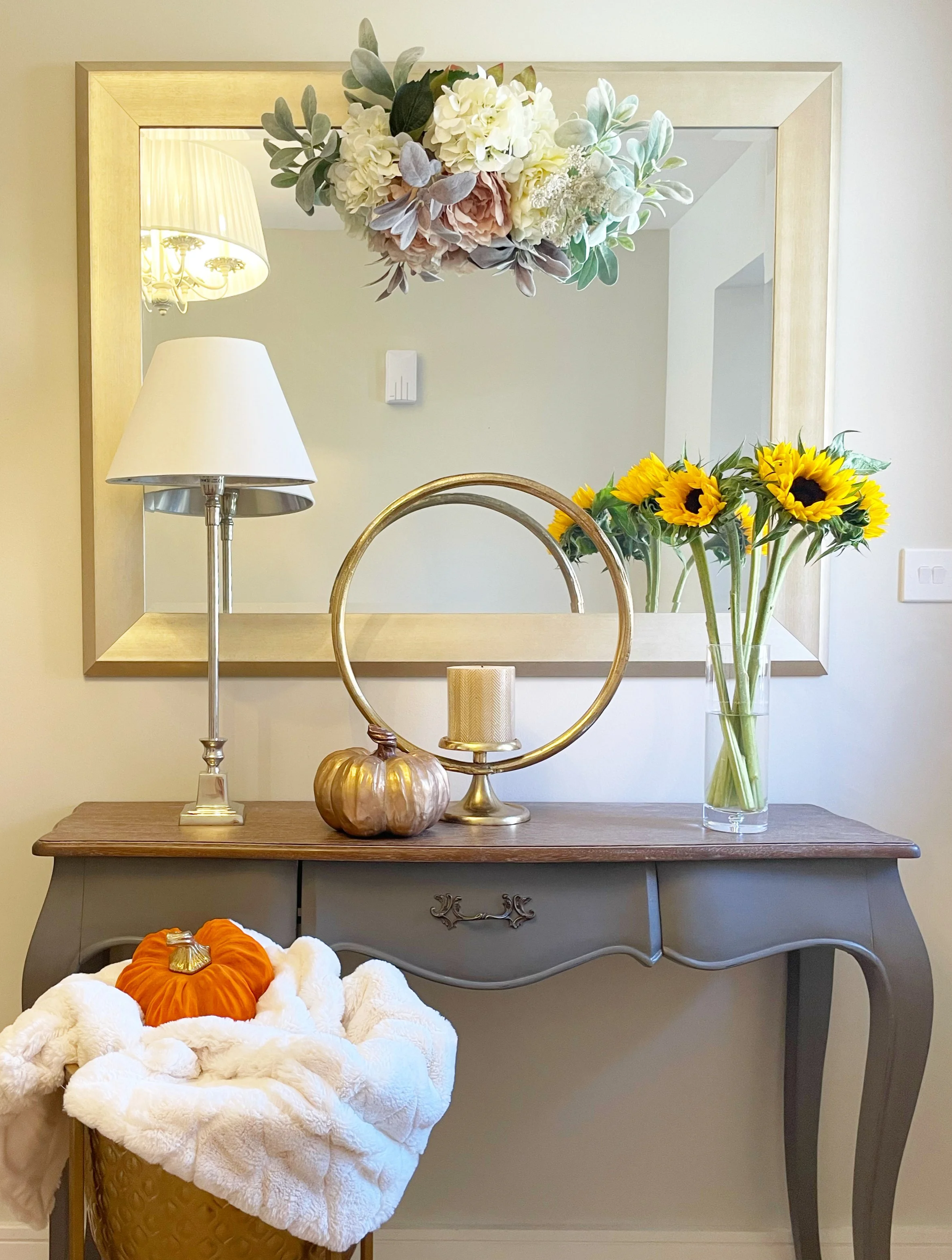 A decorative table display featuring a large mirror with a gold frame hanging on a wall. On the table, there is a tall clear glass vase with bright yellow sunflowers. To the left, there is a gold pumpkin-shaped candle holder. A small table lamp with a silver base and white shade is also on the table. In the foreground, a basket covered with white plush fabric holds a large orange pumpkin and some smaller white pumpkins.
