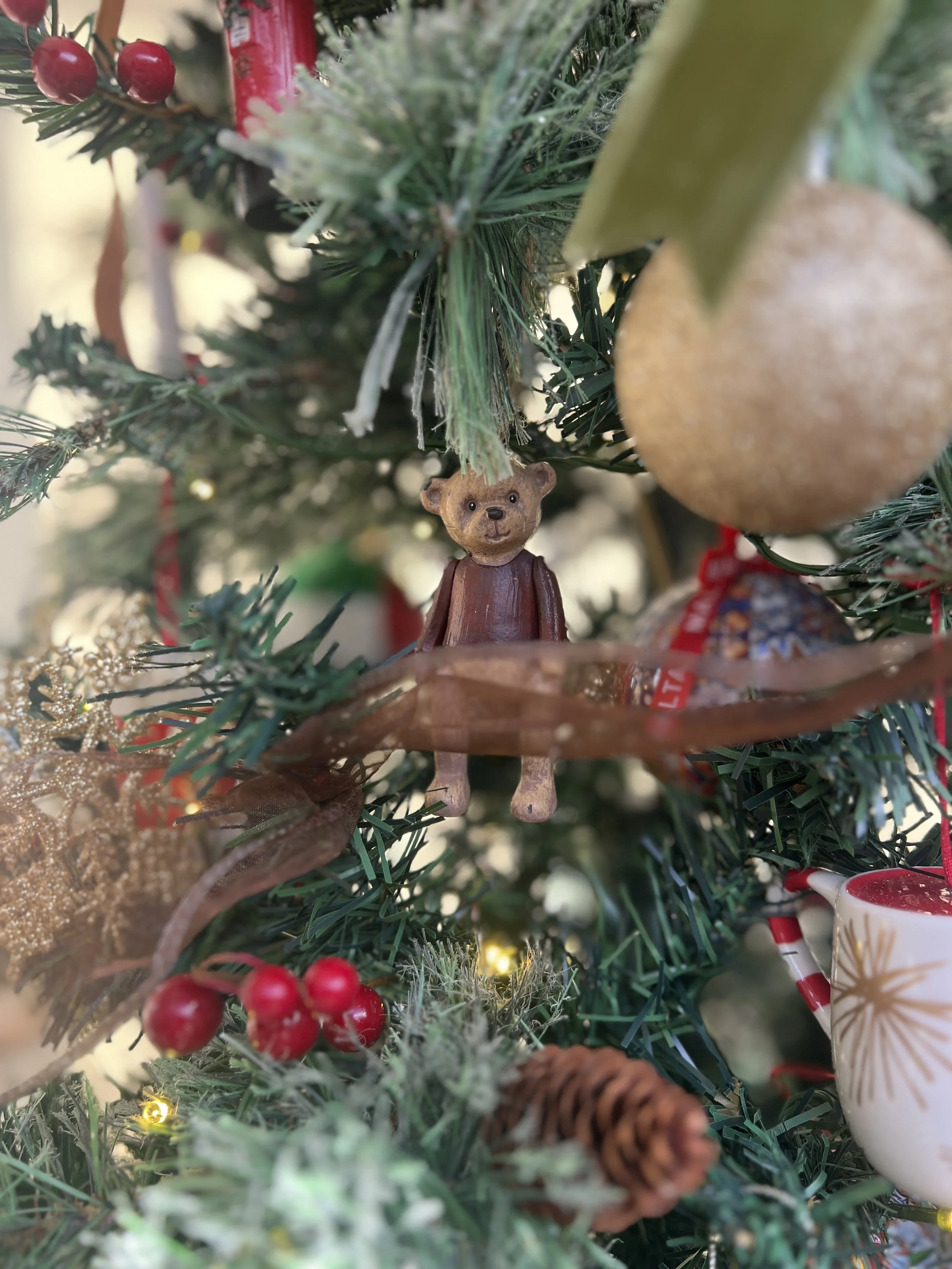 Close-up of a decorated Christmas tree with a small wooden bear ornament, red berries, a pine cone, and a gold glittery ribbon.