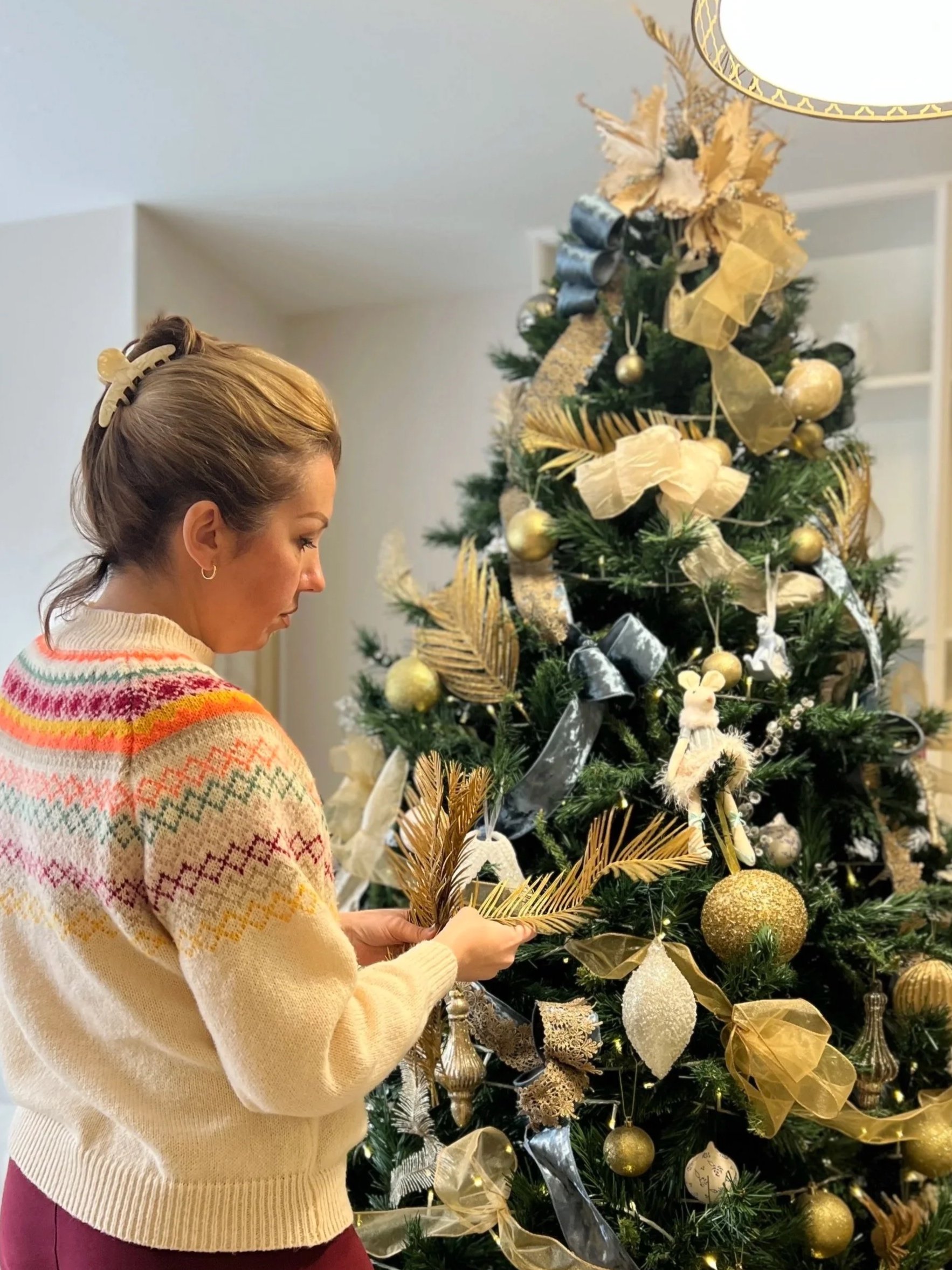 A woman in a multicolored sweater decorating a Christmas tree with gold and silver ornaments and ribbons.