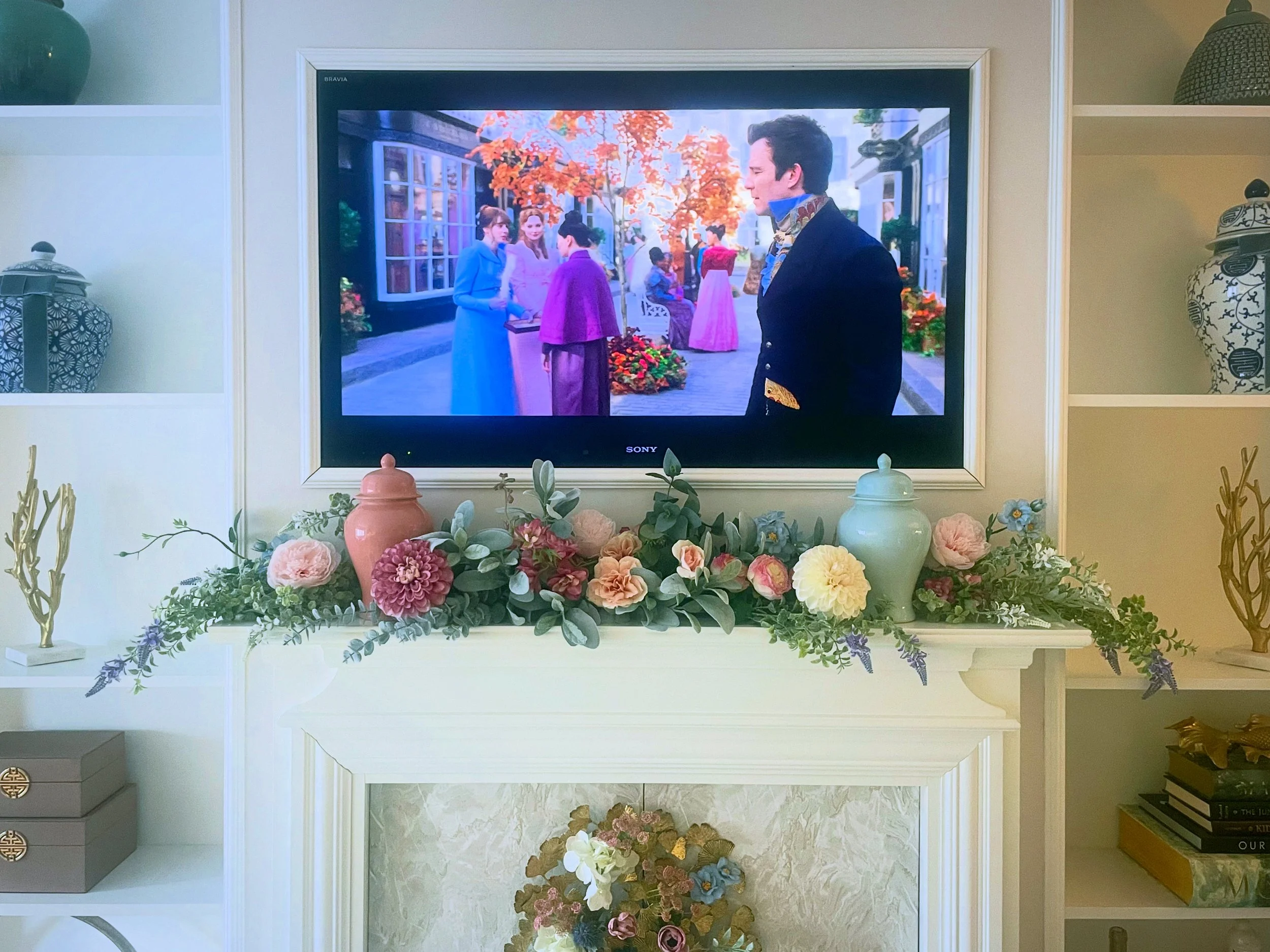 A TV displaying a scene from a period drama with people in historical costumes, surrounded by a white fireplace mantle decorated with colorful flowers and vases.
